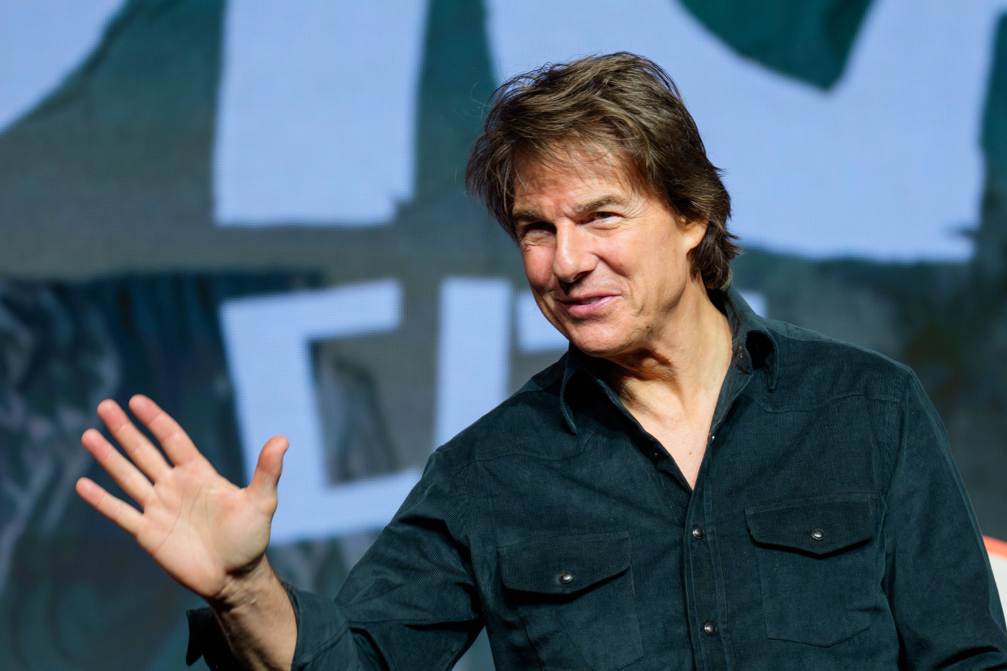 Tom Cruise in dunklem Hemd lächelt und winkt auf einer Pressekonferenz vor einem unscharfen Hintergrund.