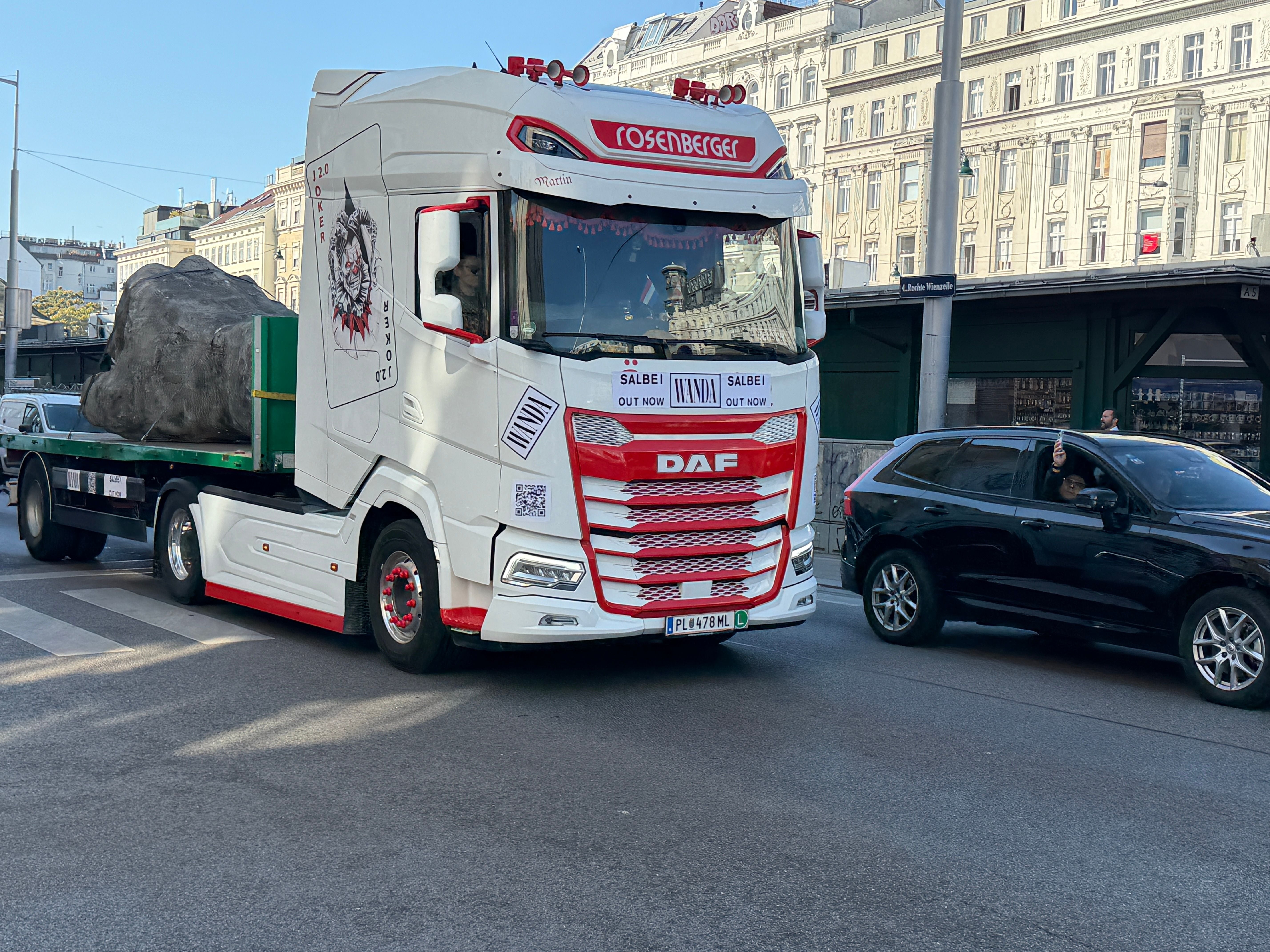 Ein weißer LKW mit großem Stein auf dem Anhänger fährt durch eine Stadtstraße.