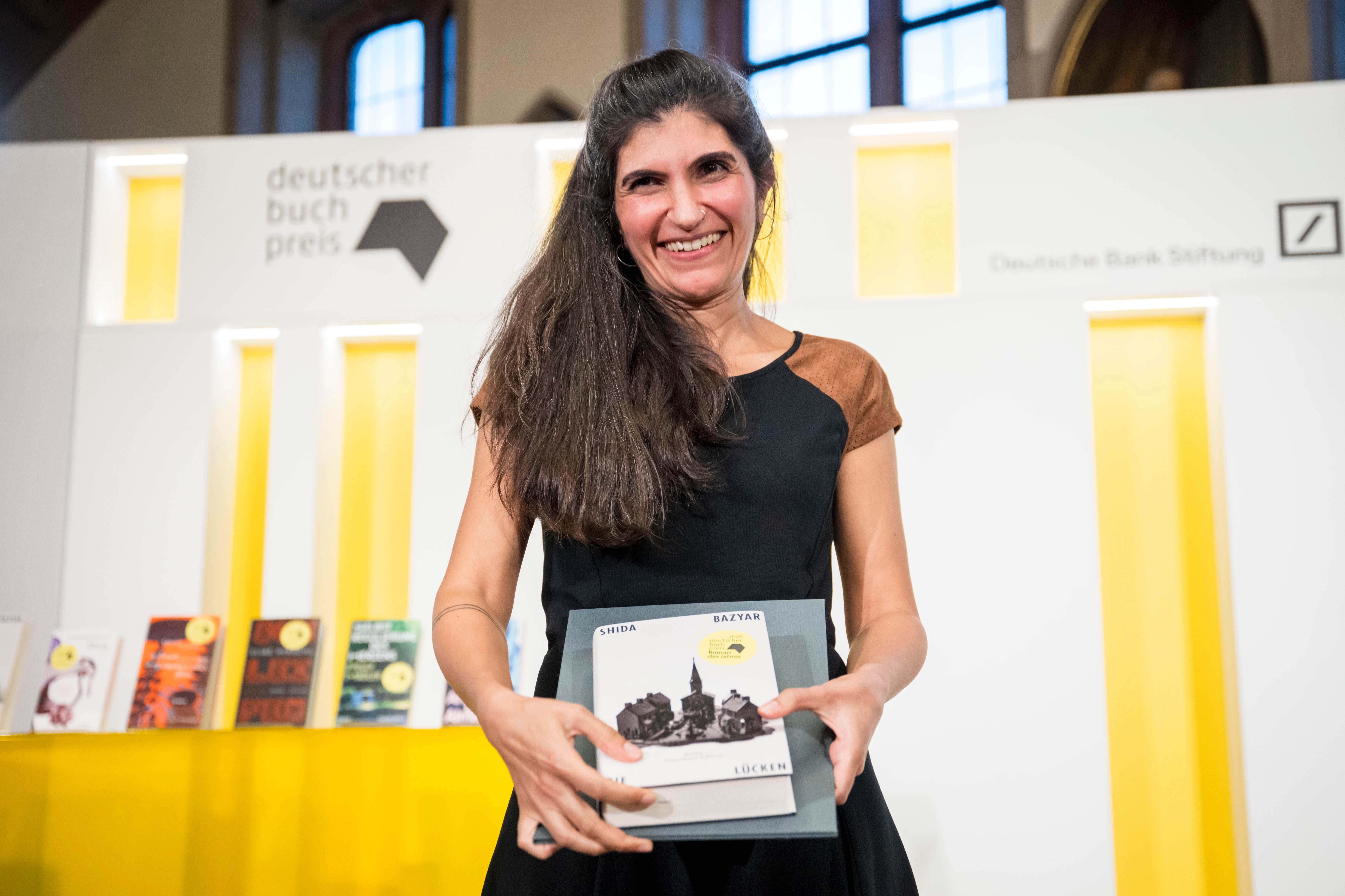 Shida Bazyar hält ihr Buch in der Hand, im Hintergrund Bücherregale und ein Schriftzug „Deutscher Buchpreis“.