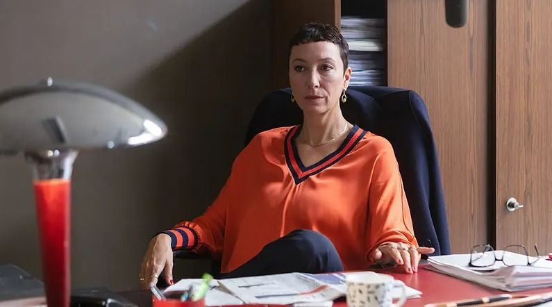 Eine Frau in orangefarbener Bluse sitzt an einem Schreibtisch in einem Büro.