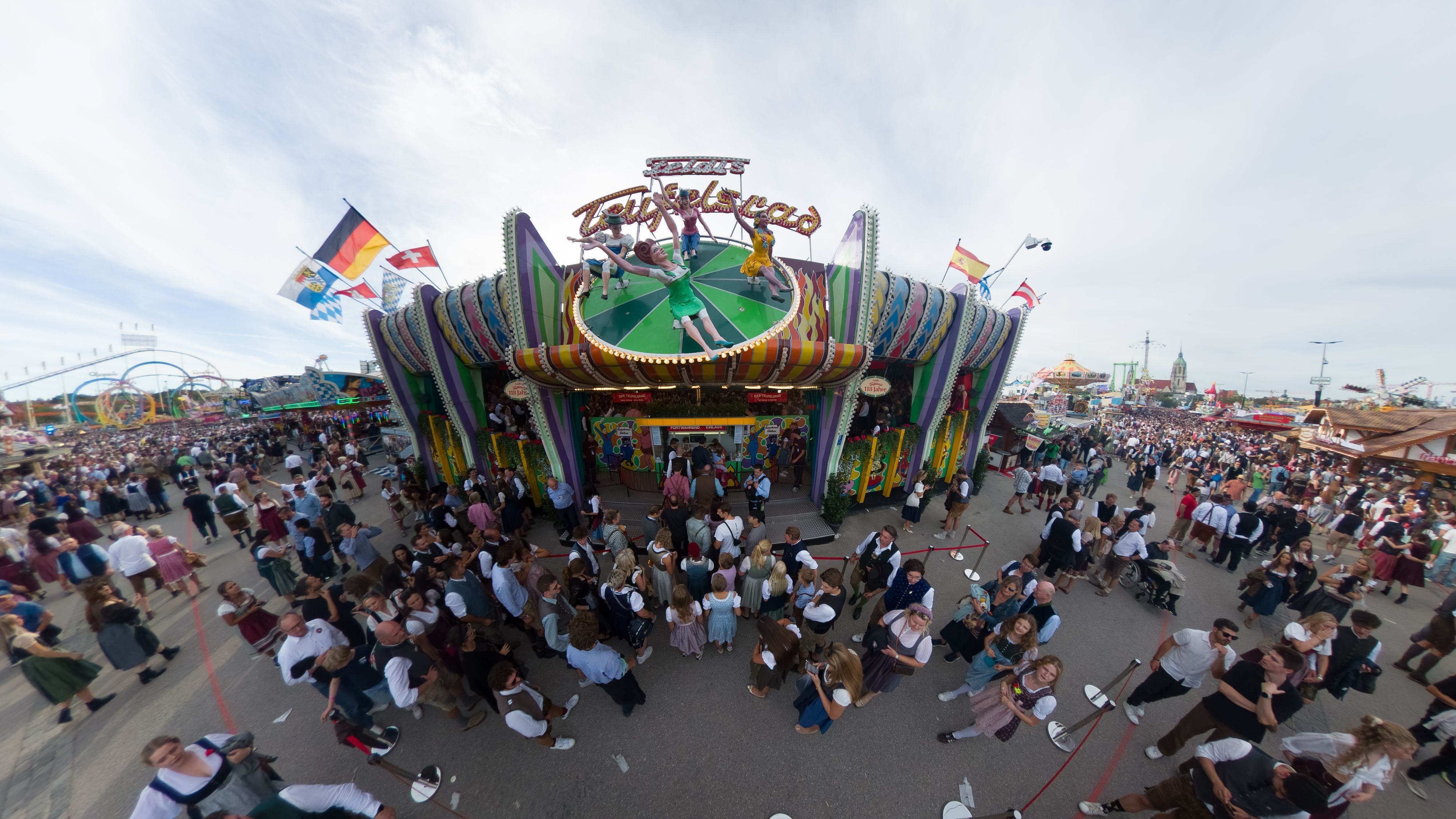 Menschenmenge vor dem Fahrgeschäft Teufelsrad auf dem Oktoberfest in München bei Tag.