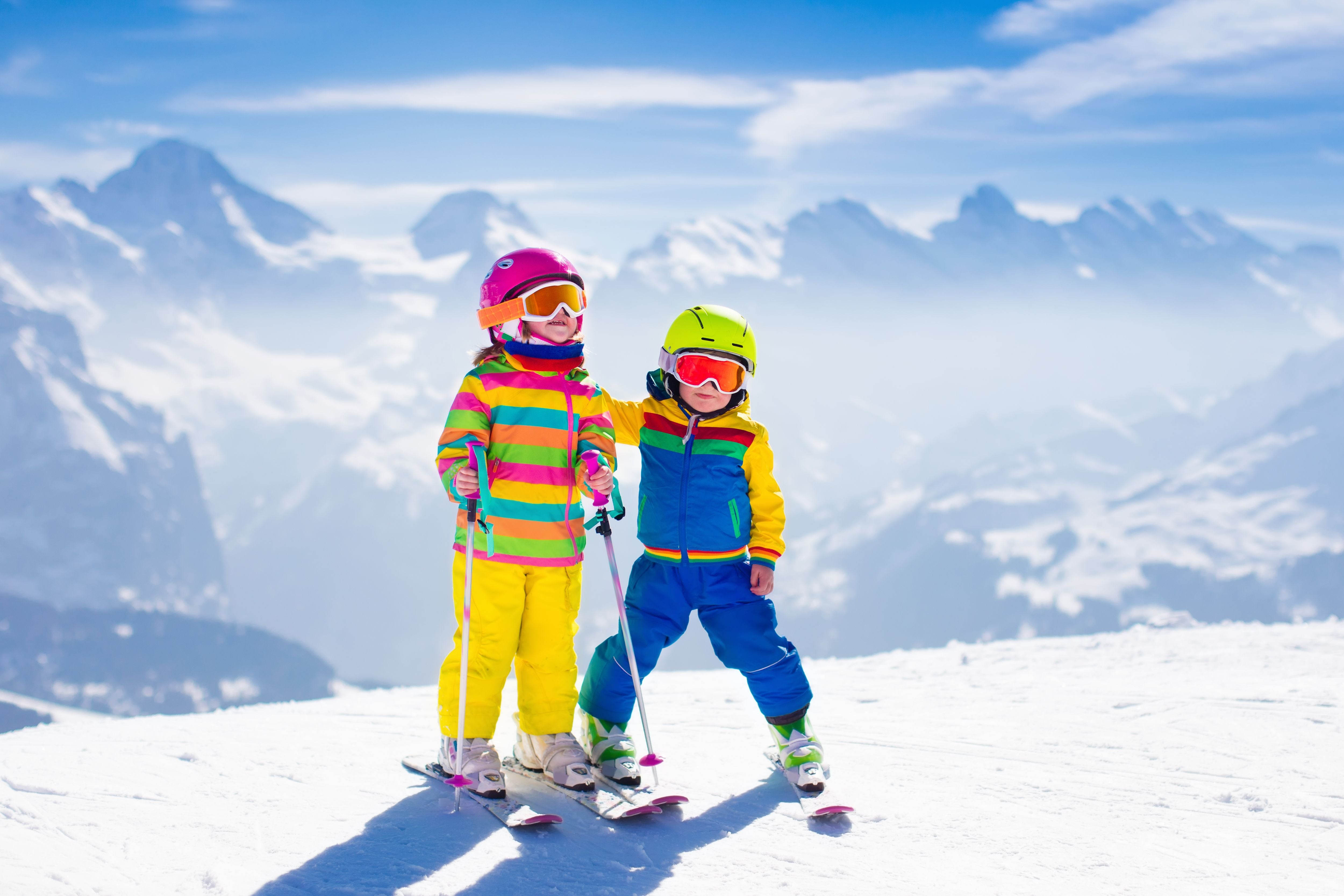 Zwei Kinder in bunter Skibekleidung stehen mit Skiern und Ausrüstung auf einer verschneiten Berglandschaft.
