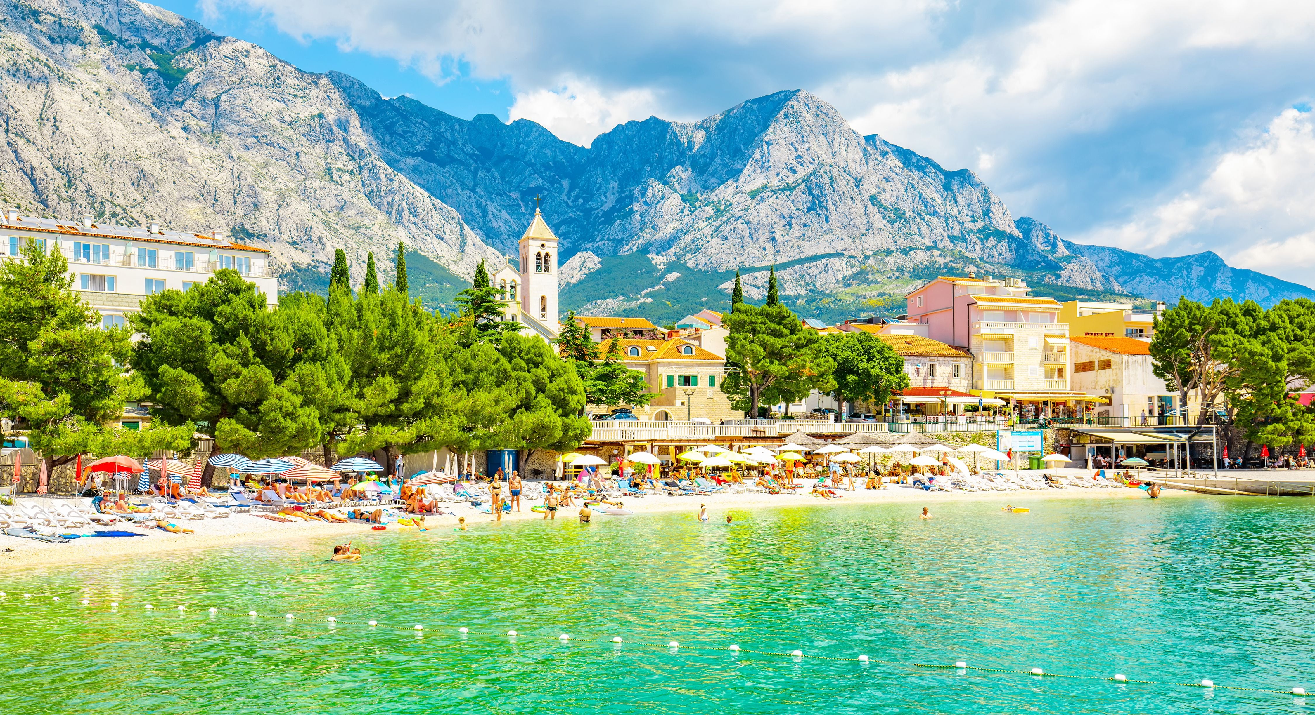 Strand in Baska Voda mit bunten Sonnenschirmen, türkisfarbenem Wasser und Bergen im Hintergrund.