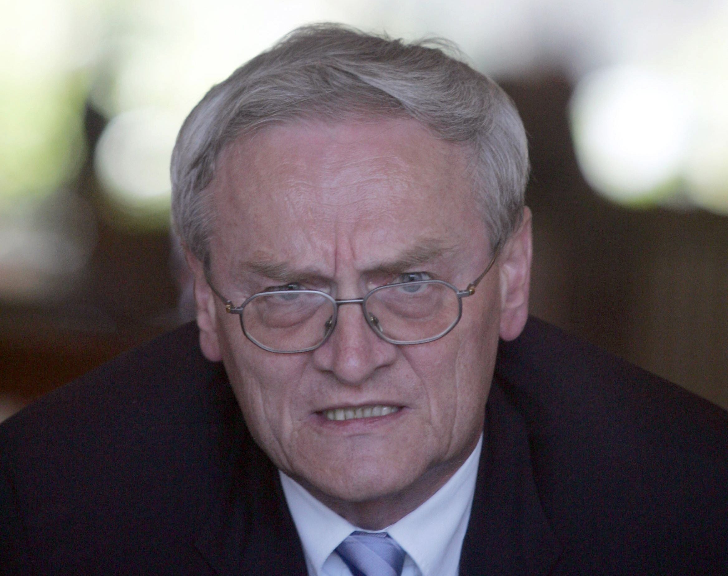 Ein älterer Mann mit Brille und Anzug blickt ernst in die Kamera.