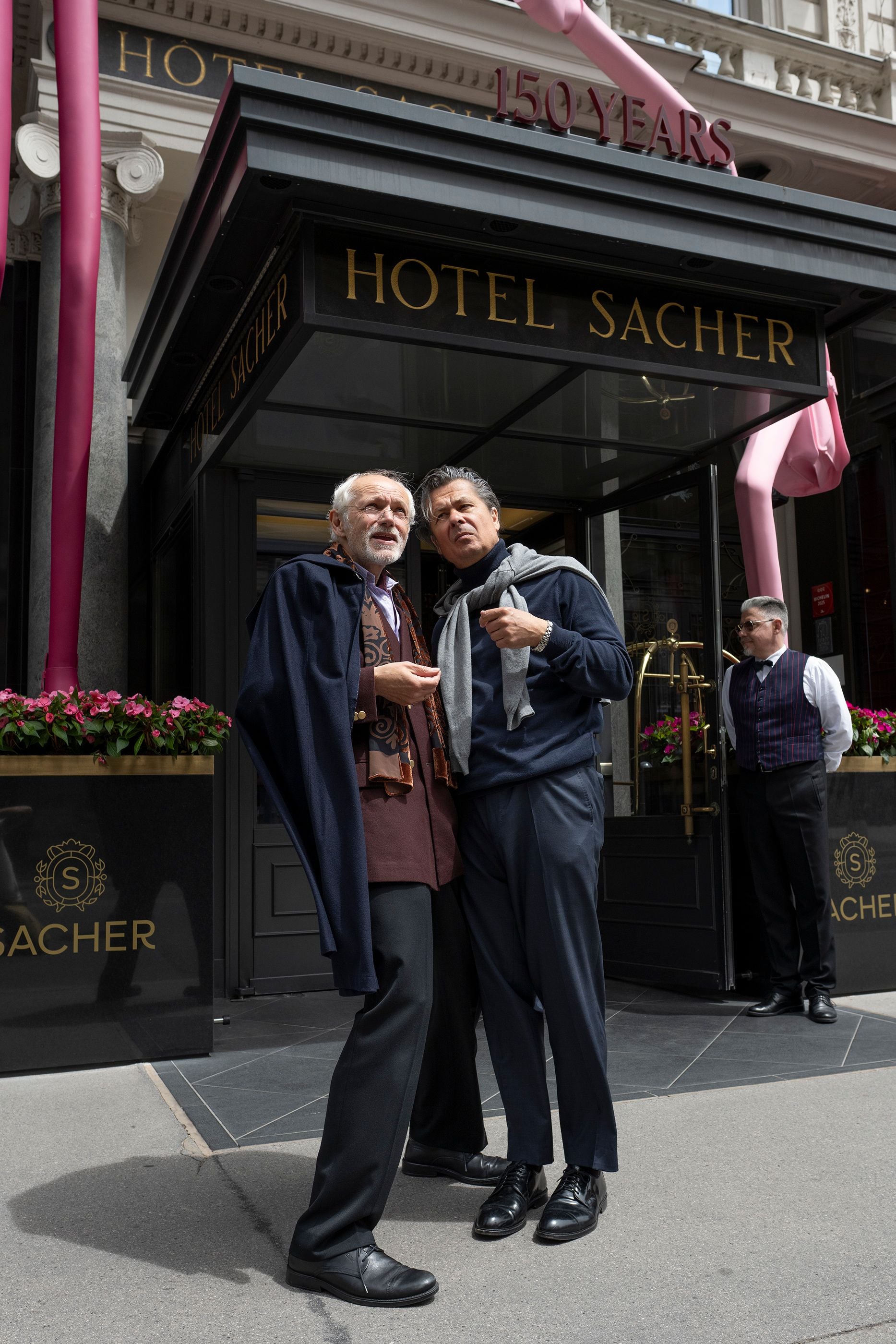 Zwei Männer stehen vor dem Hotel Sacher, im Hintergrund ein Hotelmitarbeiter und Blumenschmuck.