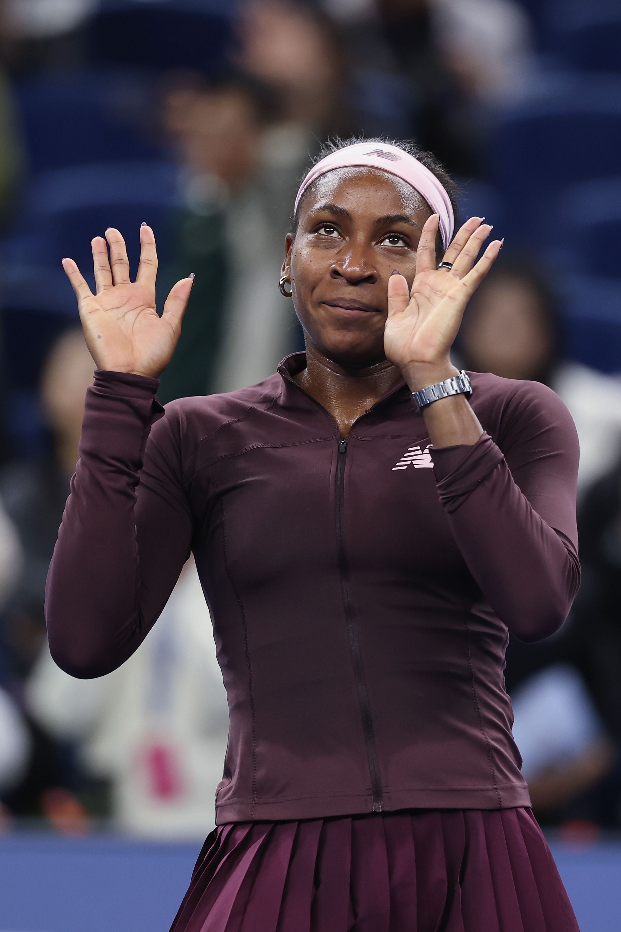 Coco Gauff hebt im dunklen Sportoberteil mit Stirnband beide Hände hoch und blickt fragend nach oben.
