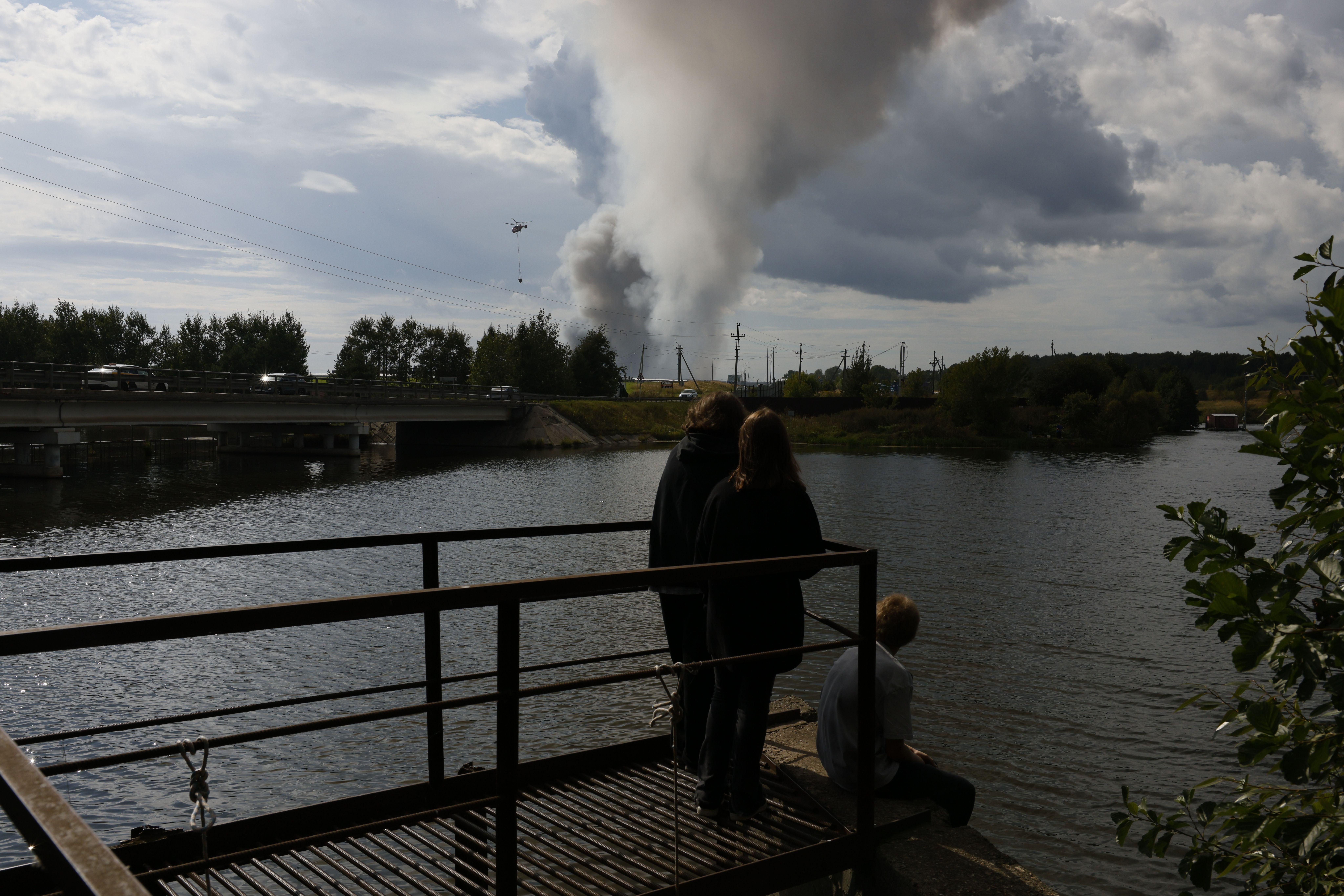 Drei Personen beobachten dichten Rauch, der von einem fernen Brand hinter einem Fluss aufsteigt.