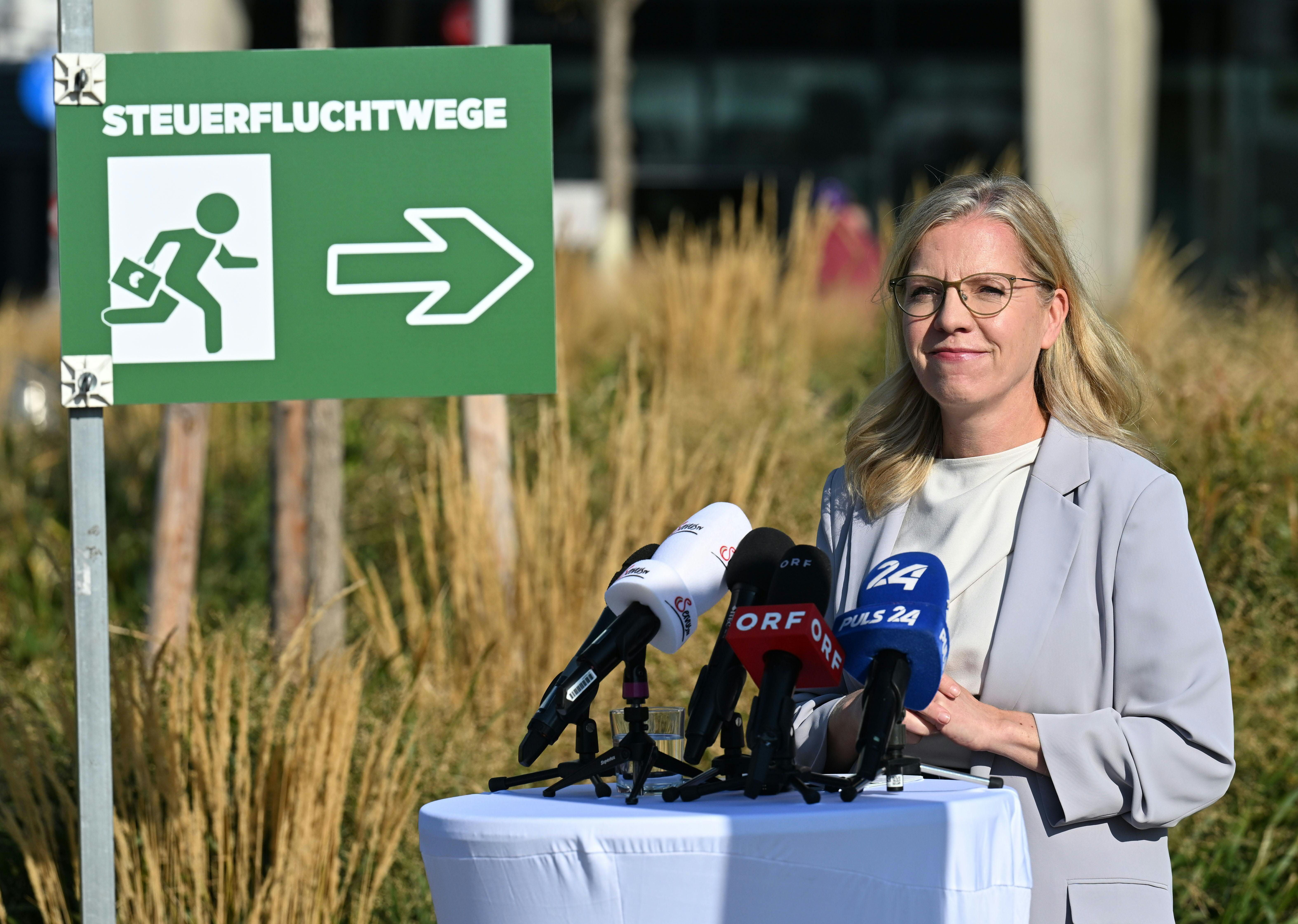 Eine Frau steht bei einer Pressekonferenz mit Mikrofonen neben einem Schild "STEUERFLUCHTWEGE".