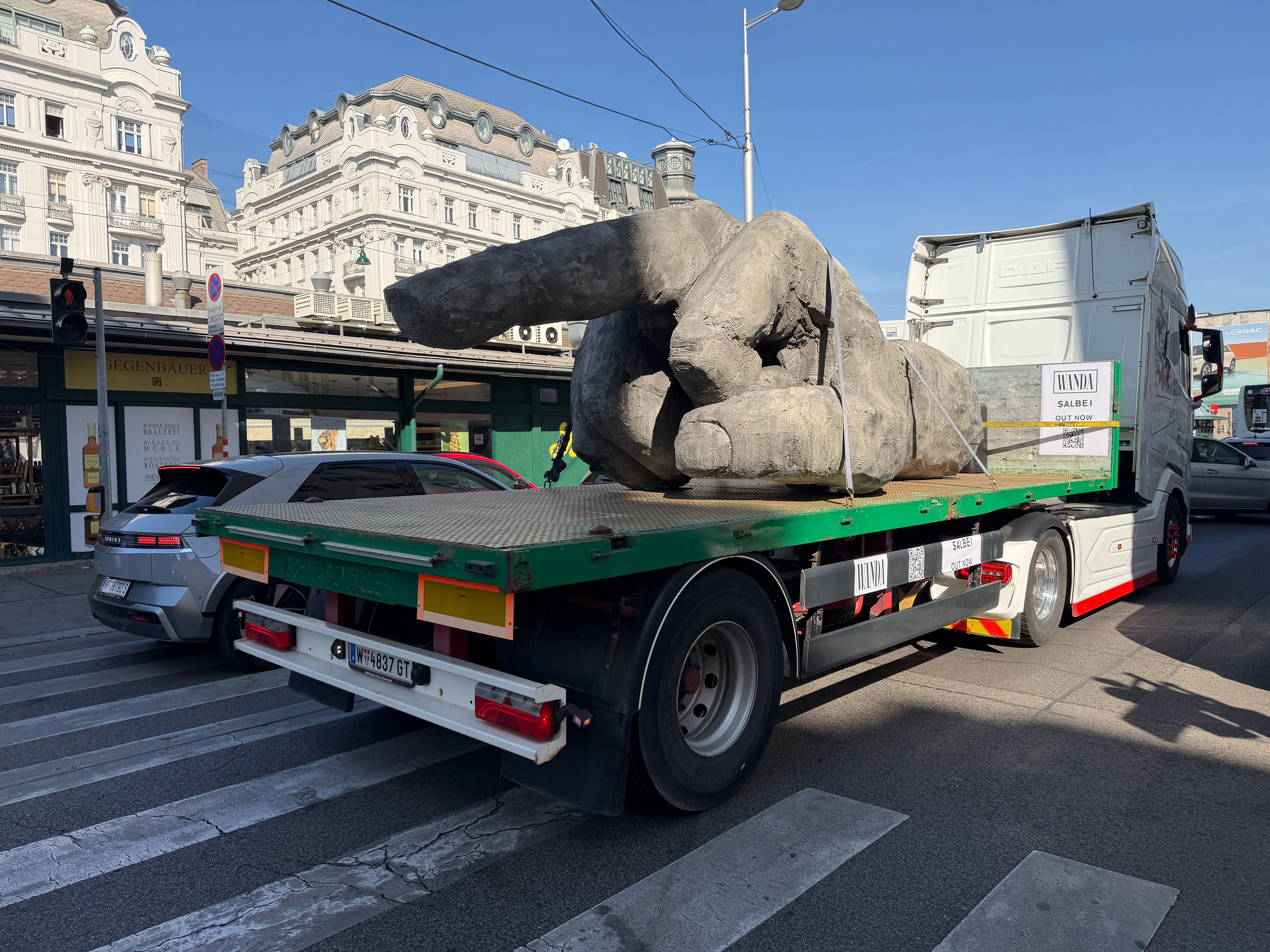 Ein LKW transportiert eine große Skulptur einer Hand durch eine Stadt mit historischen Gebäuden.