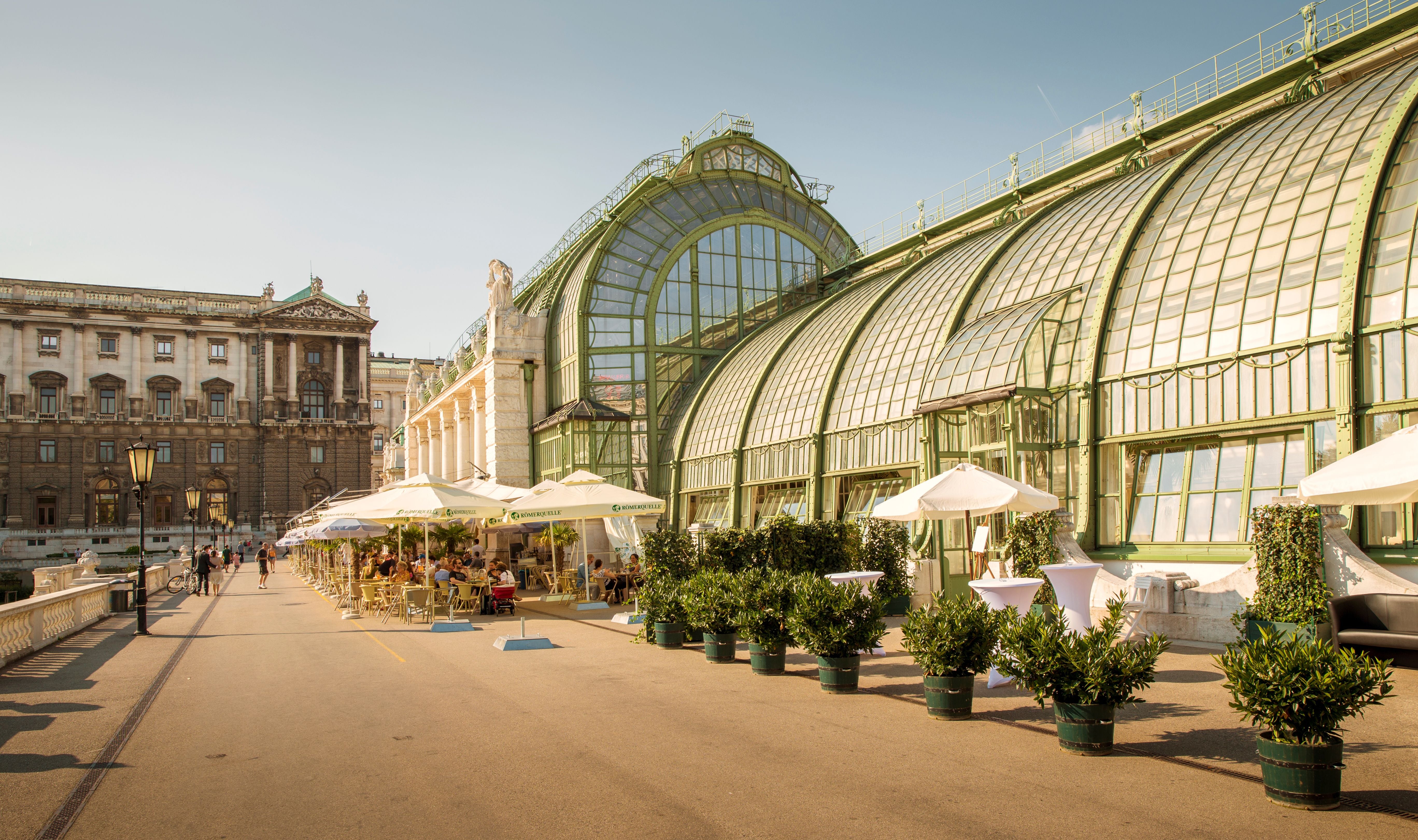 Palmenhaus in Wien mit Außencafé, Sonnenschirmen und grünen Pflanzen bei sonnigem Wetter.