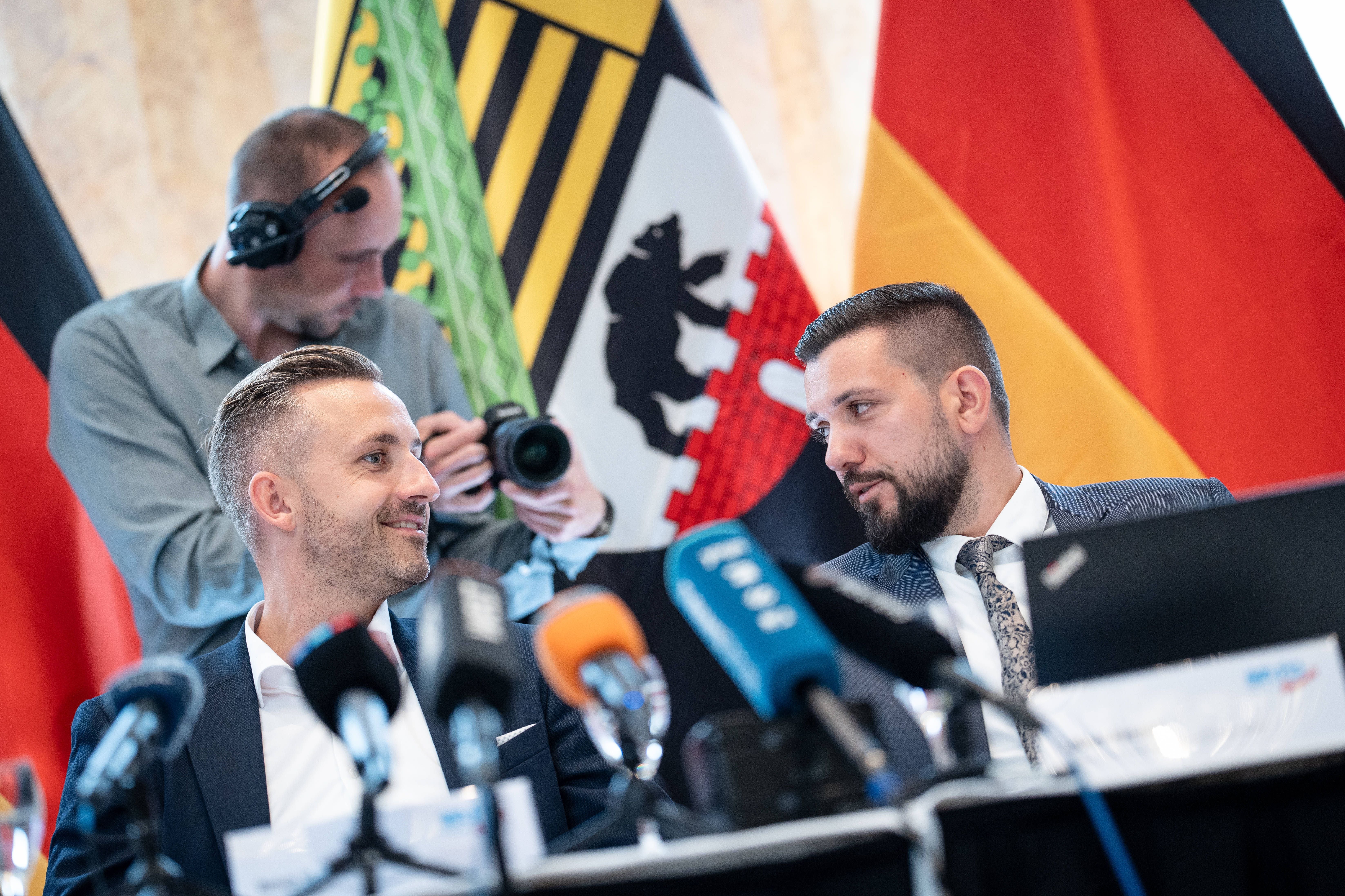 Zwei Männer im Anzug sprechen bei einer Pressekonferenz vor Mikrofonen, im Hintergrund Flaggen.