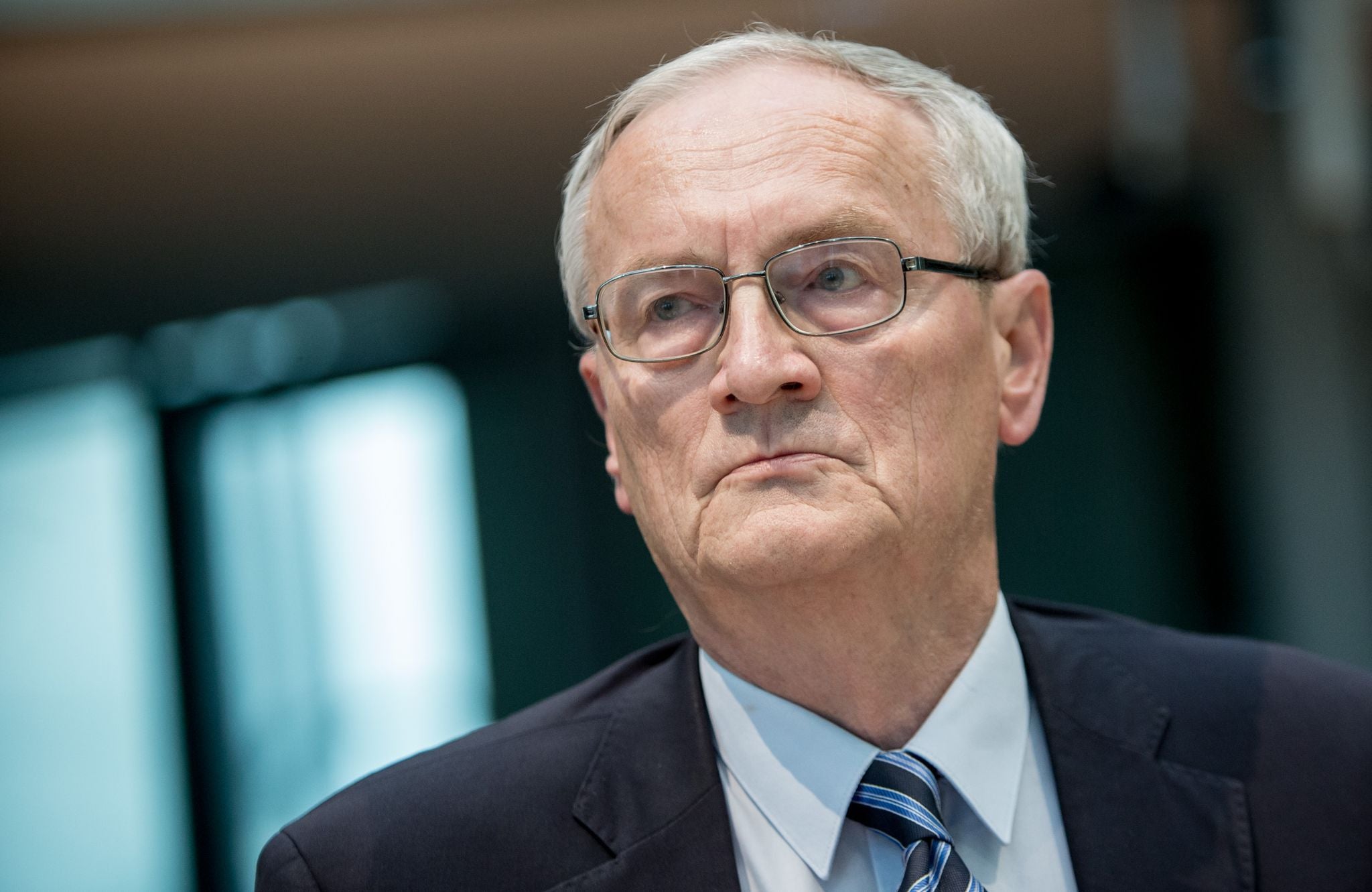 Ein älterer Mann mit Brille, Anzug und Krawatte schaut ernsthaft nach vorne.