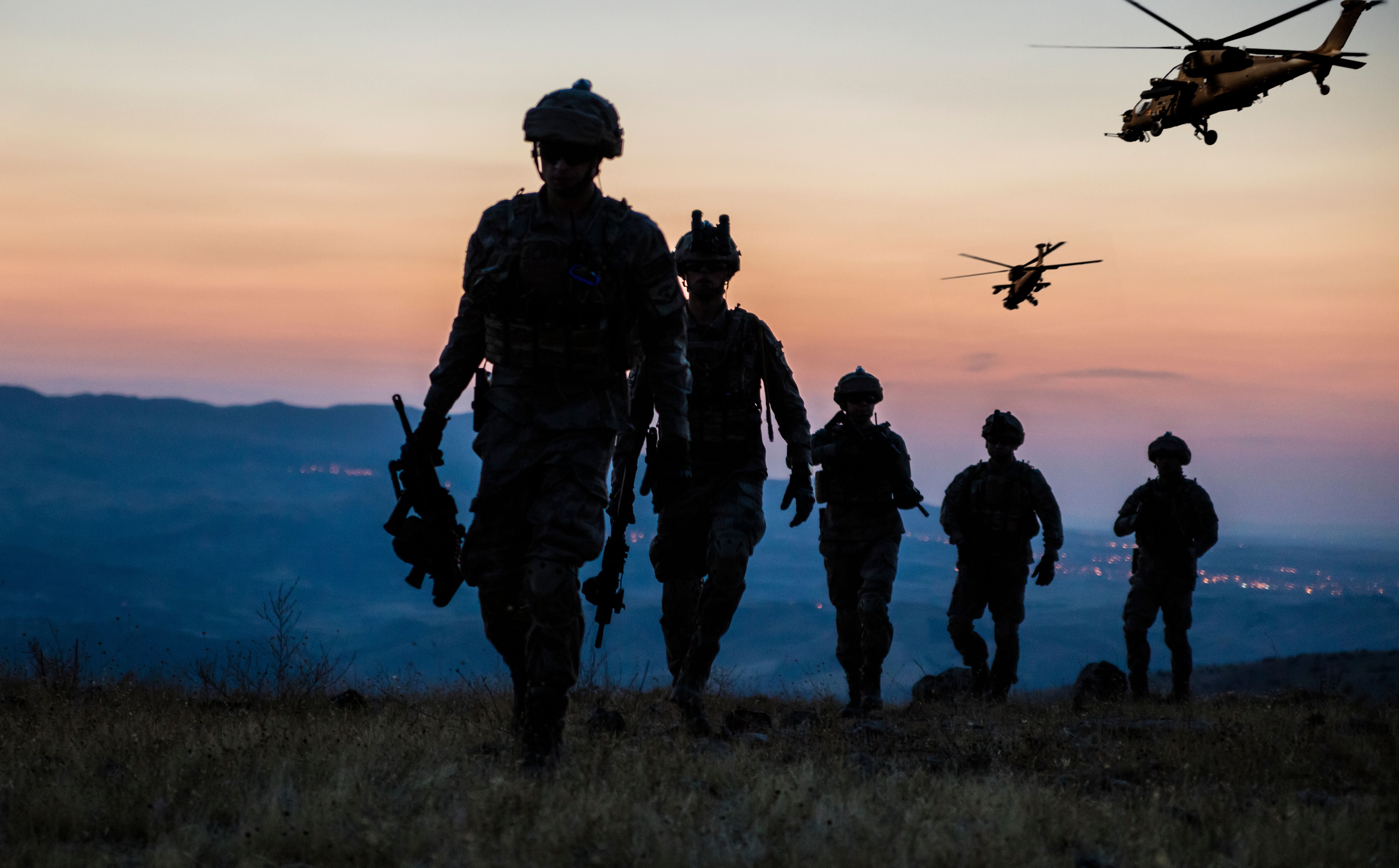 Fünf Soldaten gehen bei Dämmerung über ein Feld, zwei Hubschrauber fliegen im Hintergrund.