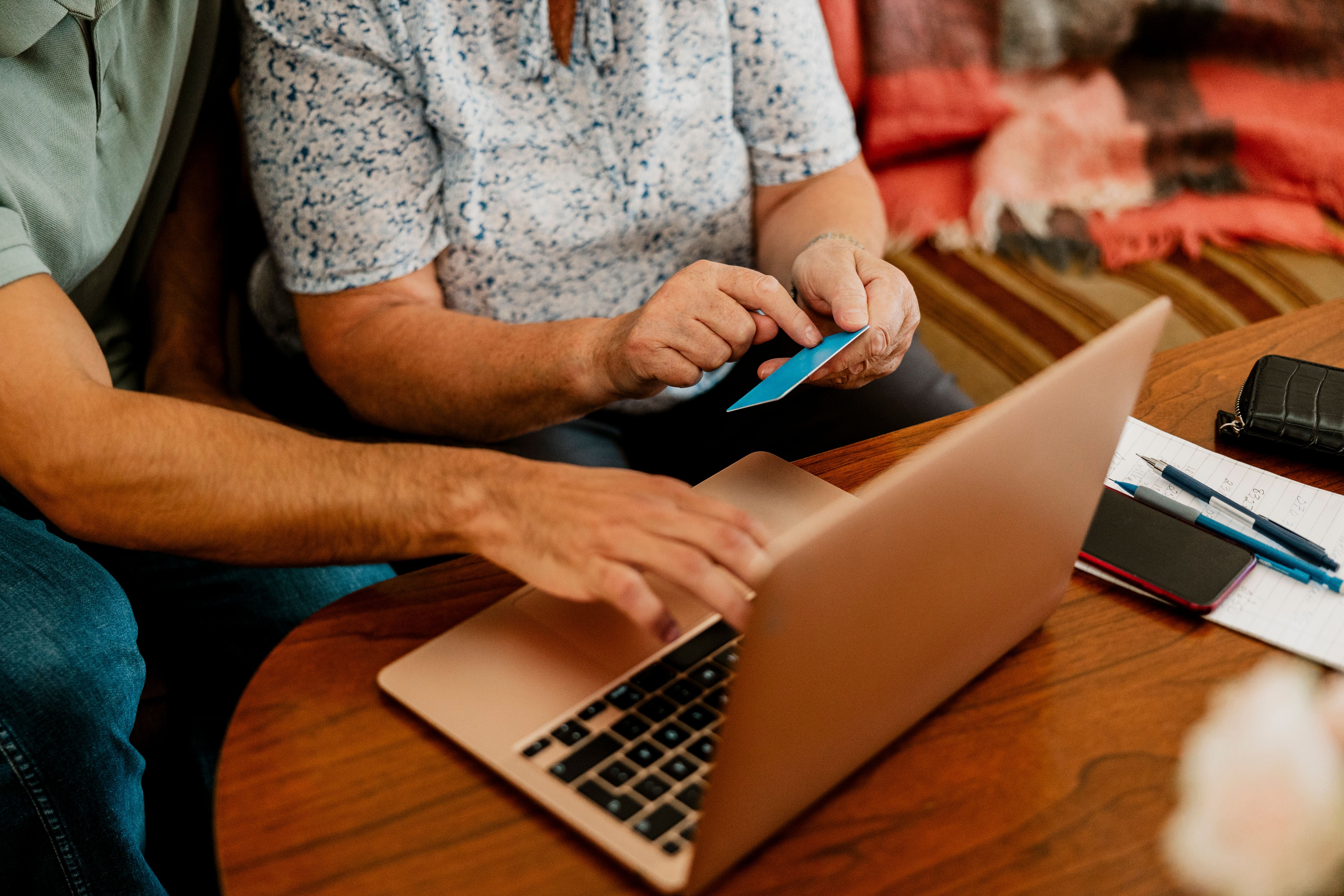 Zwei Personen benutzen einen Laptop für Online-Banking mit einer Bankkarte am Holztisch.