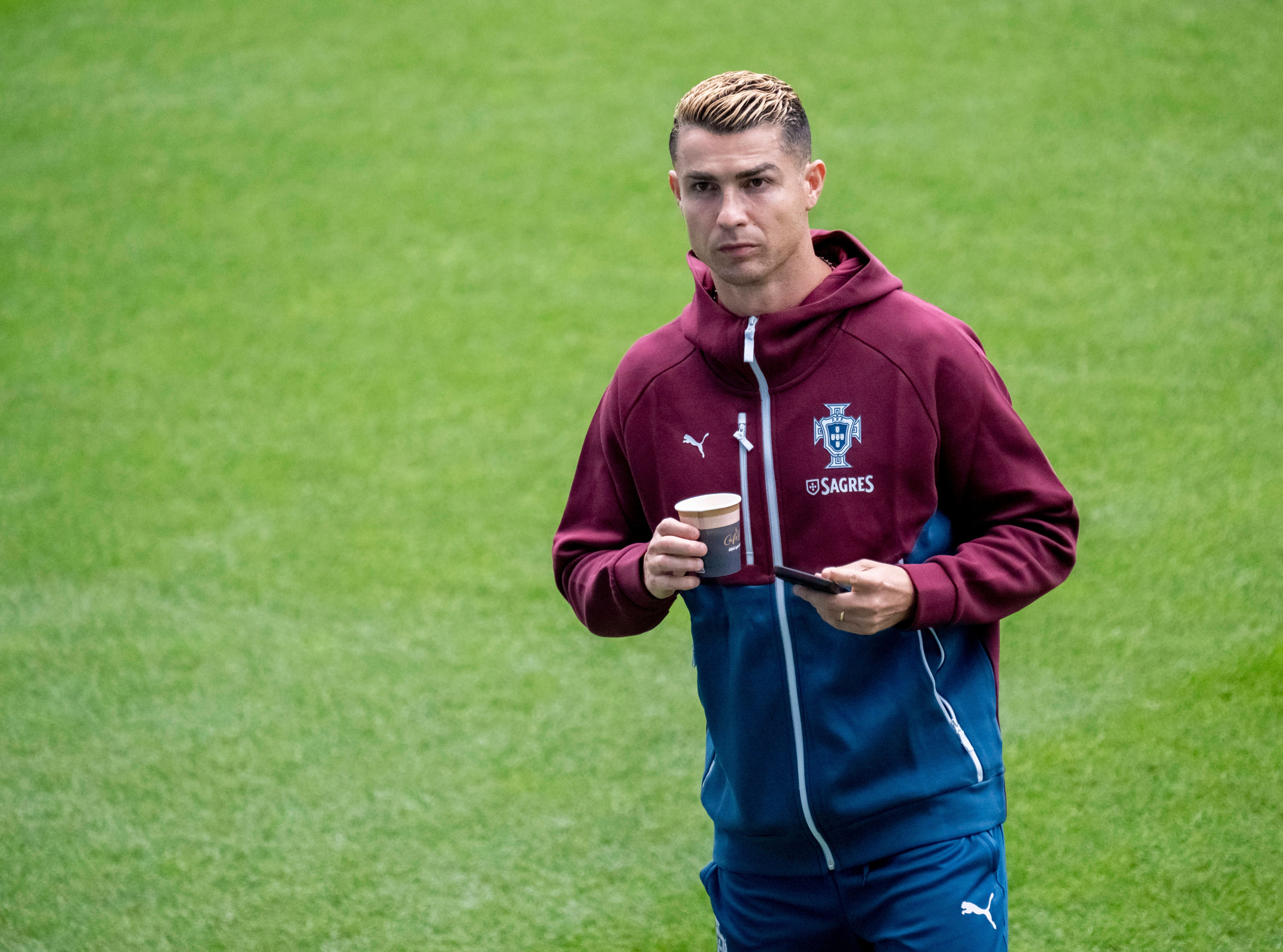 Ein Fußballspieler im Portugal-Trainingsanzug hält einen Kaffeebecher und ein Handy auf einem Rasen.