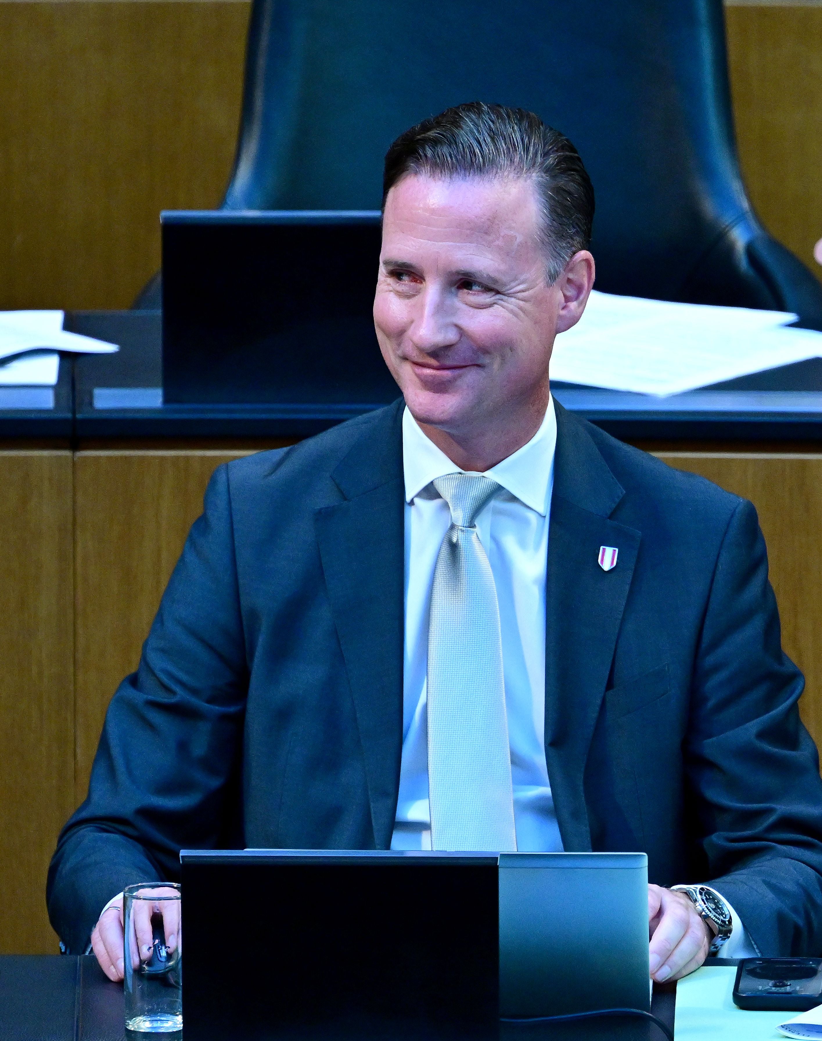 Ein Mann im Anzug sitzt lächelnd an einem Schreibtisch mit Laptop und Papier im Parlament.