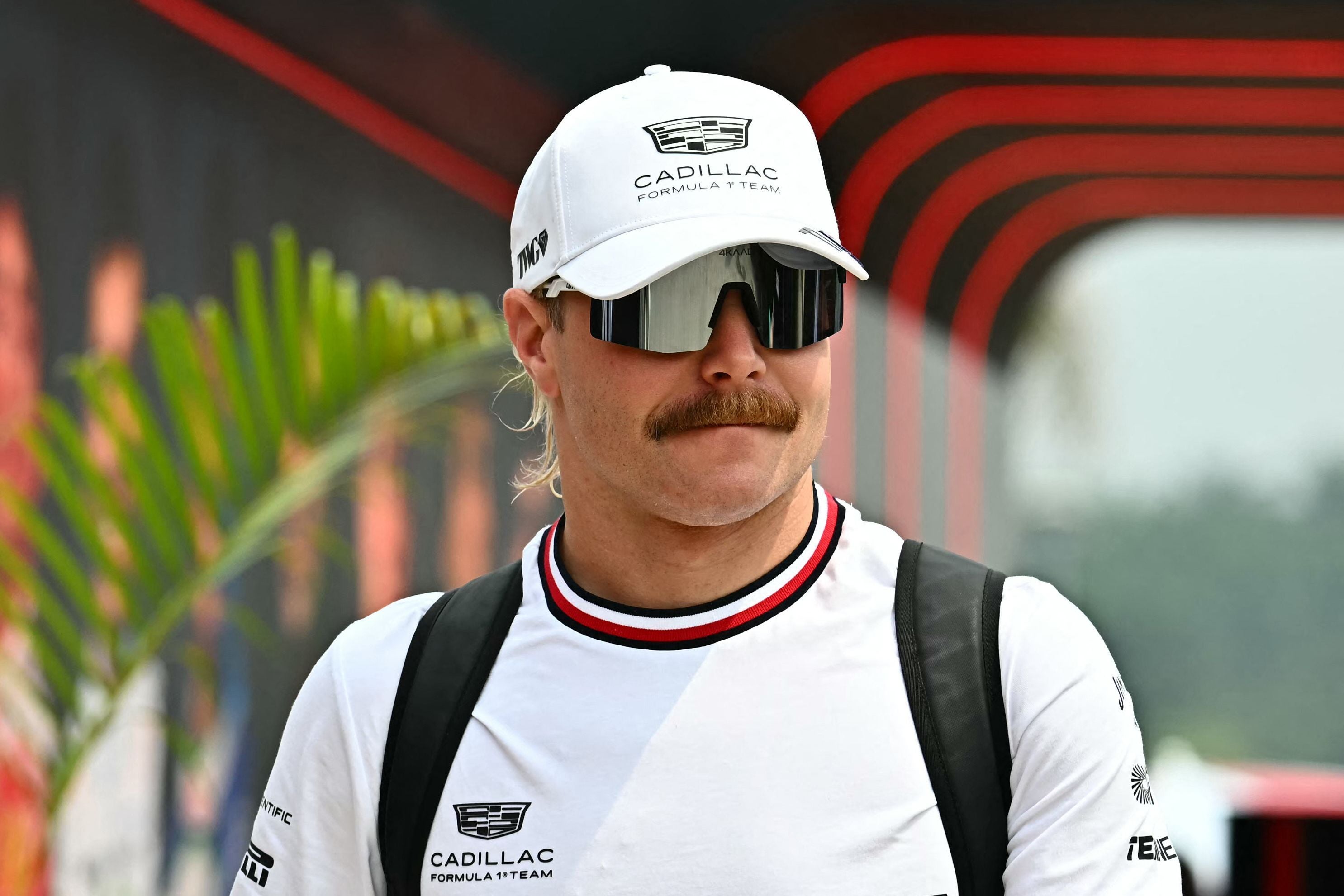 Ein Mann mit Schnurrbart, Sonnenbrille und weißem Cadillac-Basecap geht vor einer Rennstrecke.