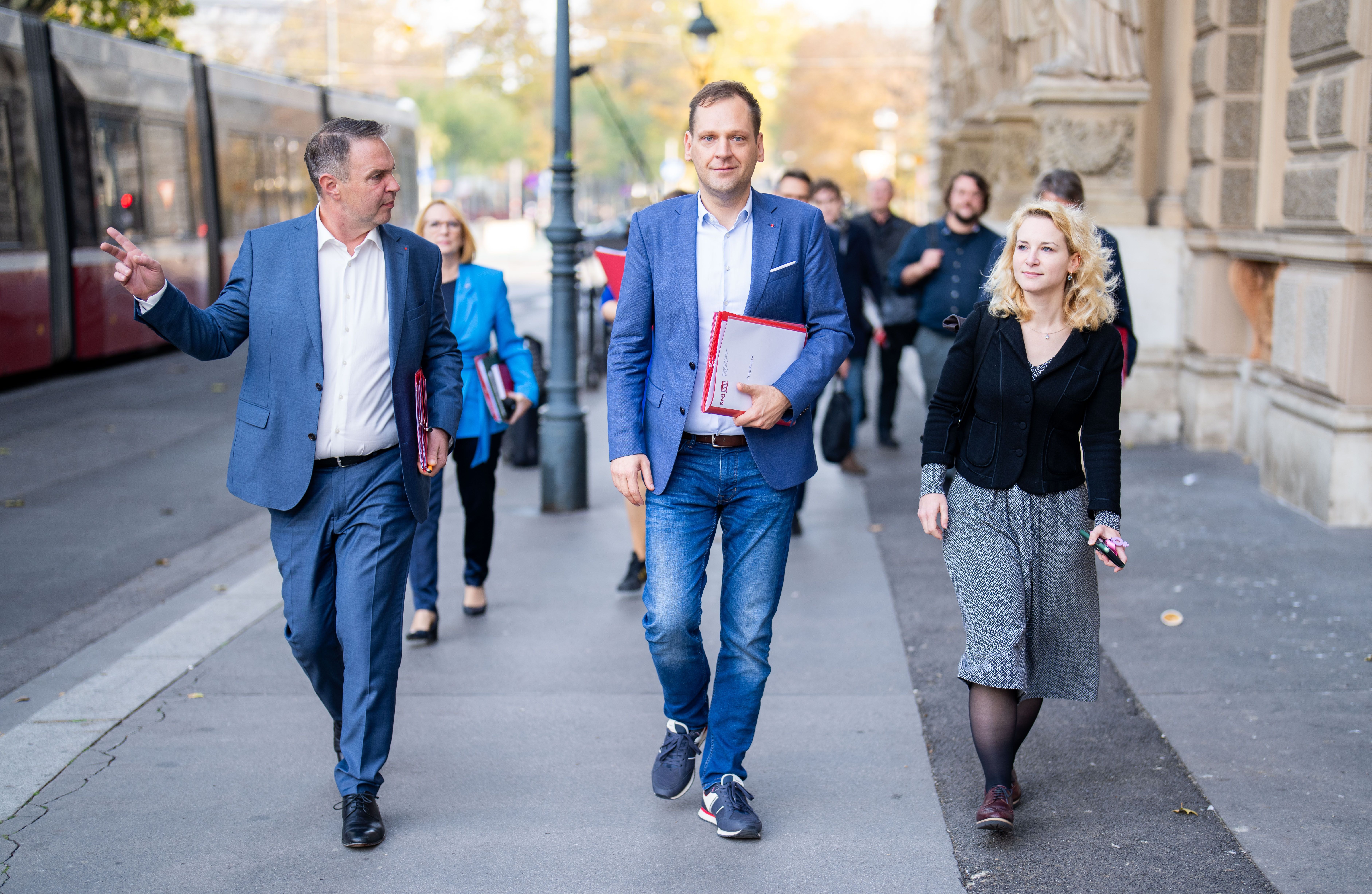 Mehrere Menschen gehen auf einem Gehweg in einer Stadt, drei vorne in Businesskleidung.
