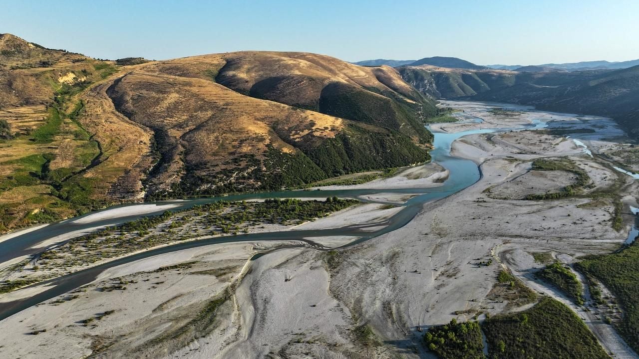 Breiter Fluss mit Kiesbänken in einer bergigen, teils bewaldeten Landschaft bei klarem Wetter.