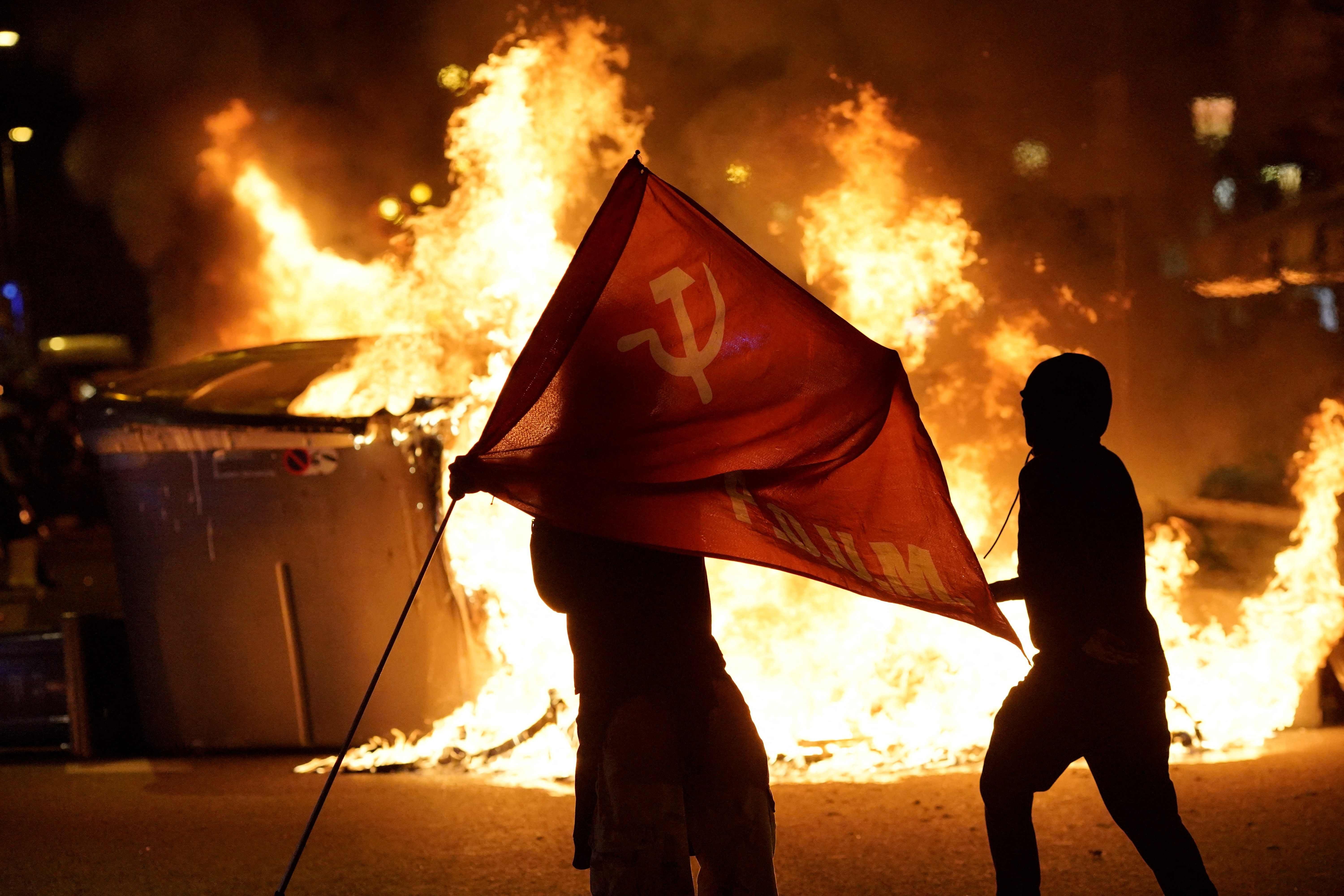 Zwei Personen mit kommunistischer Flagge stehen vor brennender Mülltonne bei einer nächtlichen Demonstration.