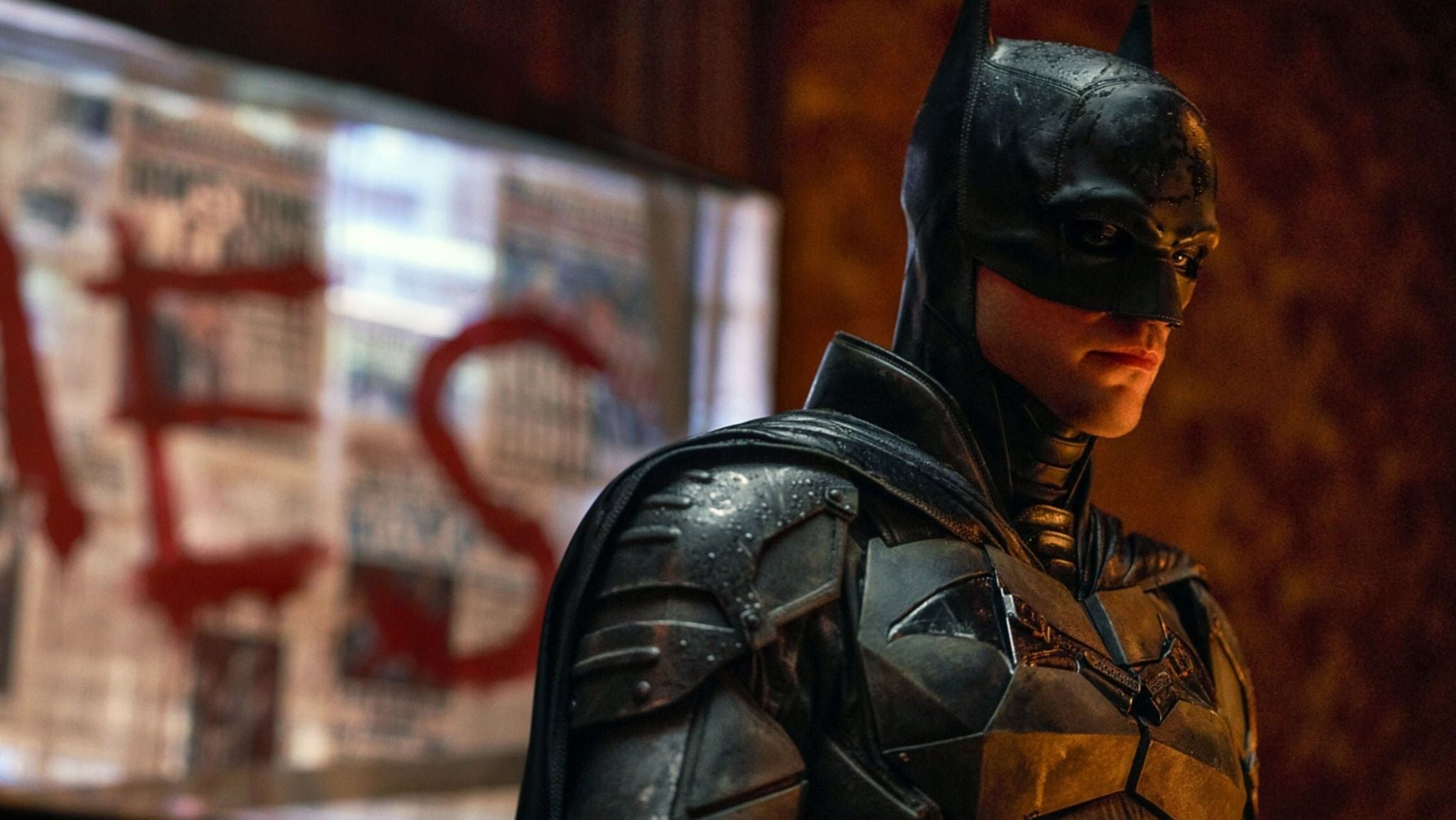 Batman steht in dunkler Rüstung vor einer Wand mit Zeitungen und roter Schrift.