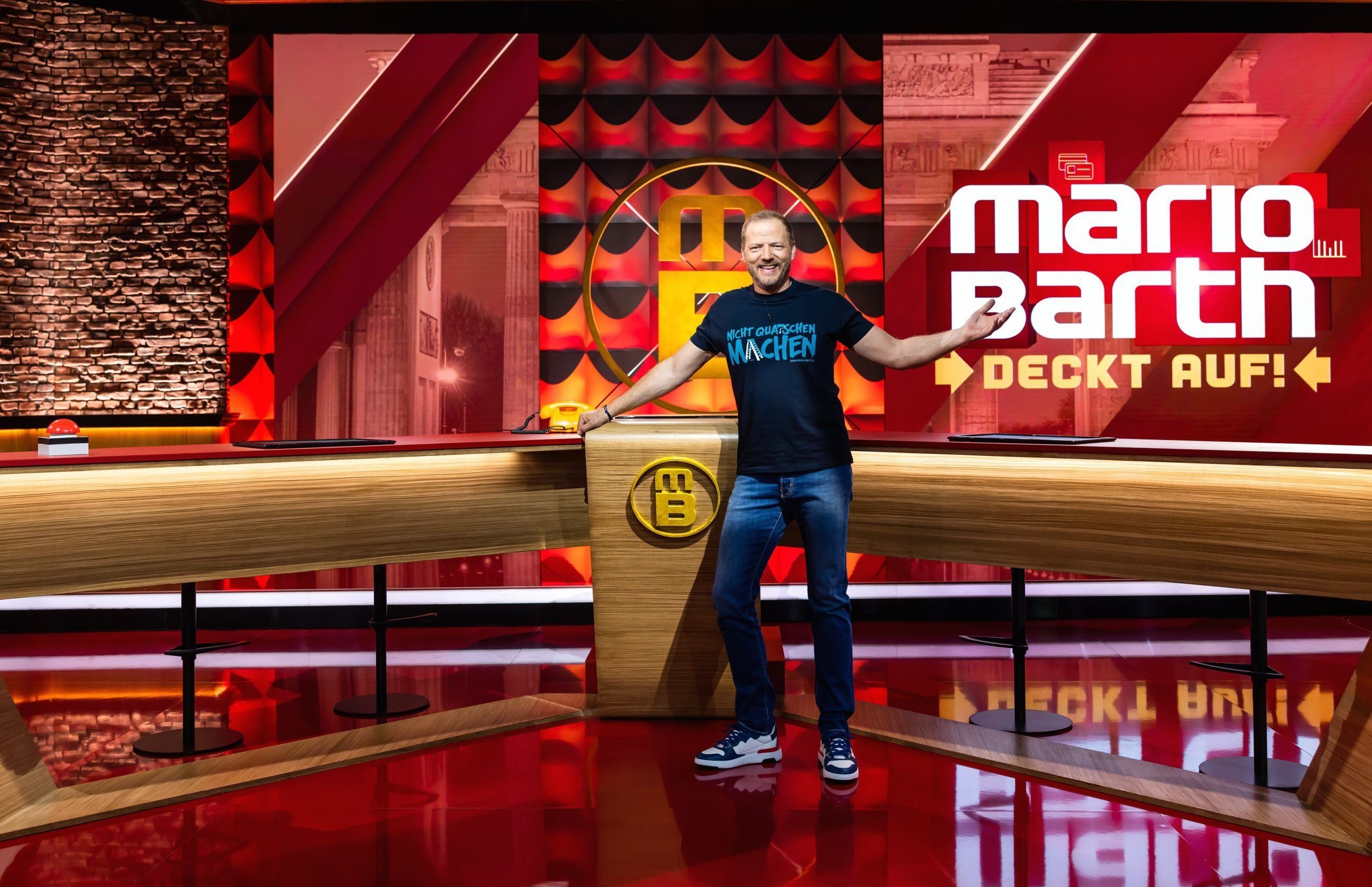 Ein Mann steht lächelnd vor dem TV-Show-Logo "Mario Barth deckt auf!" in einem modernen Studio.