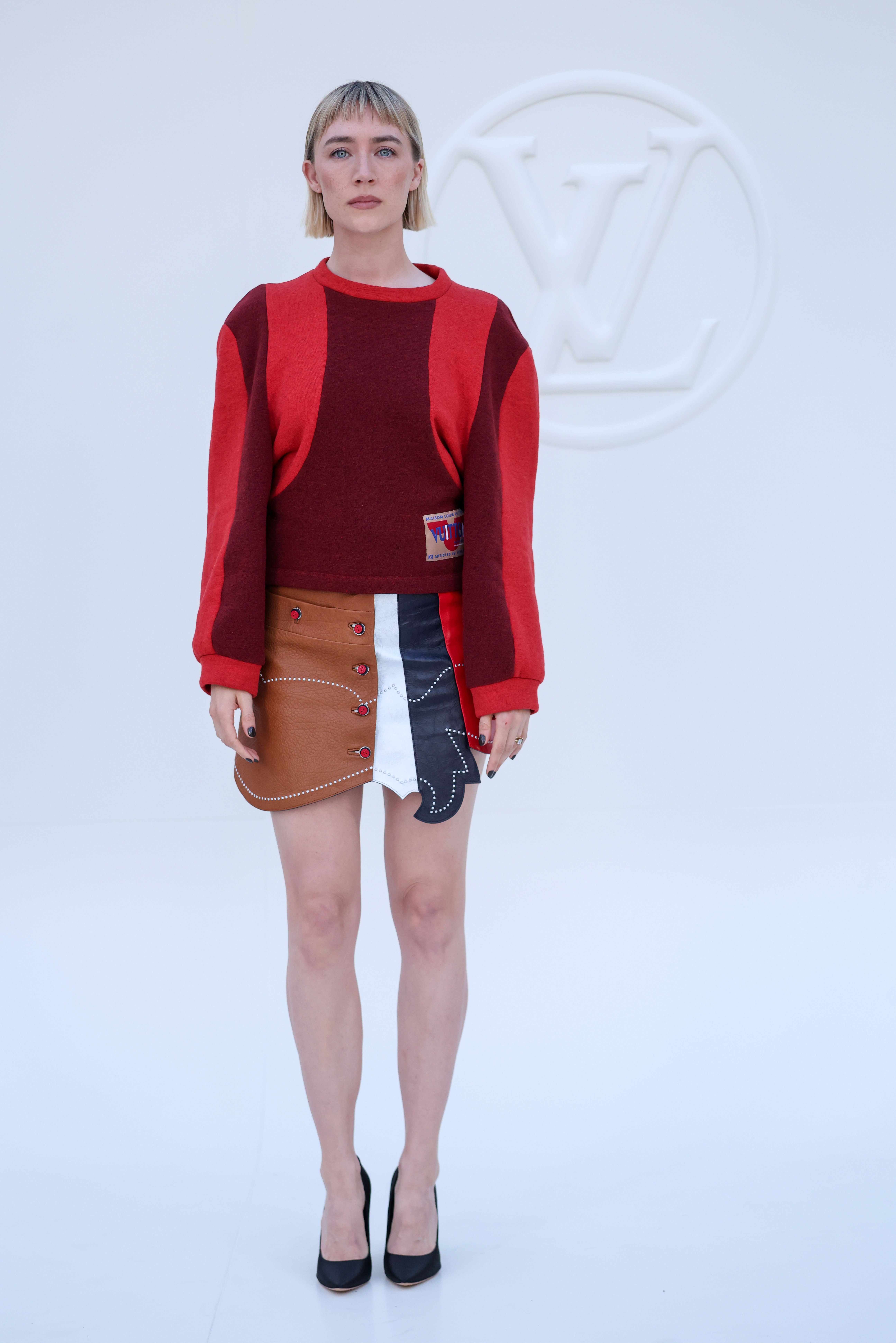 Frau in rotem Pullover und farbiger Rock steht vor Louis Vuitton Logo auf weißem Hintergrund.