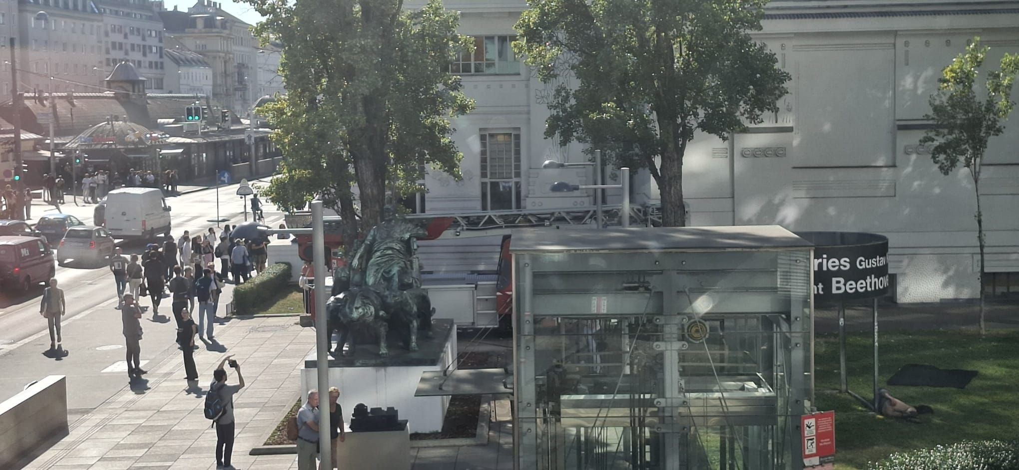 Stadtansicht mit Fußgängern, einer Pferdeskulptur und modernen Glaskonstruktionen.