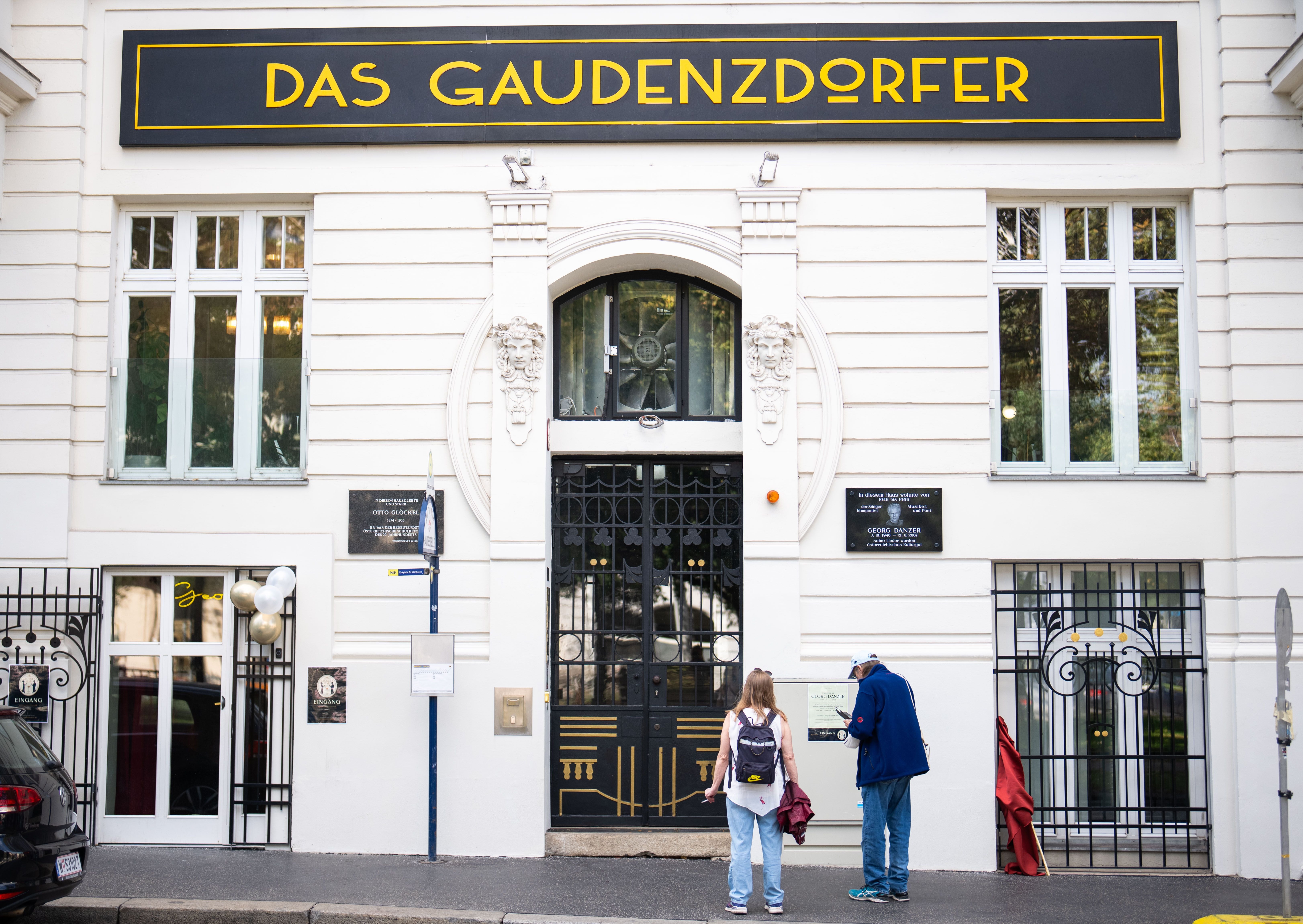 Zwei Personen stehen vor dem Gebäude "Das Gaudenzdorfer" mit Gedenktafel in Wien.