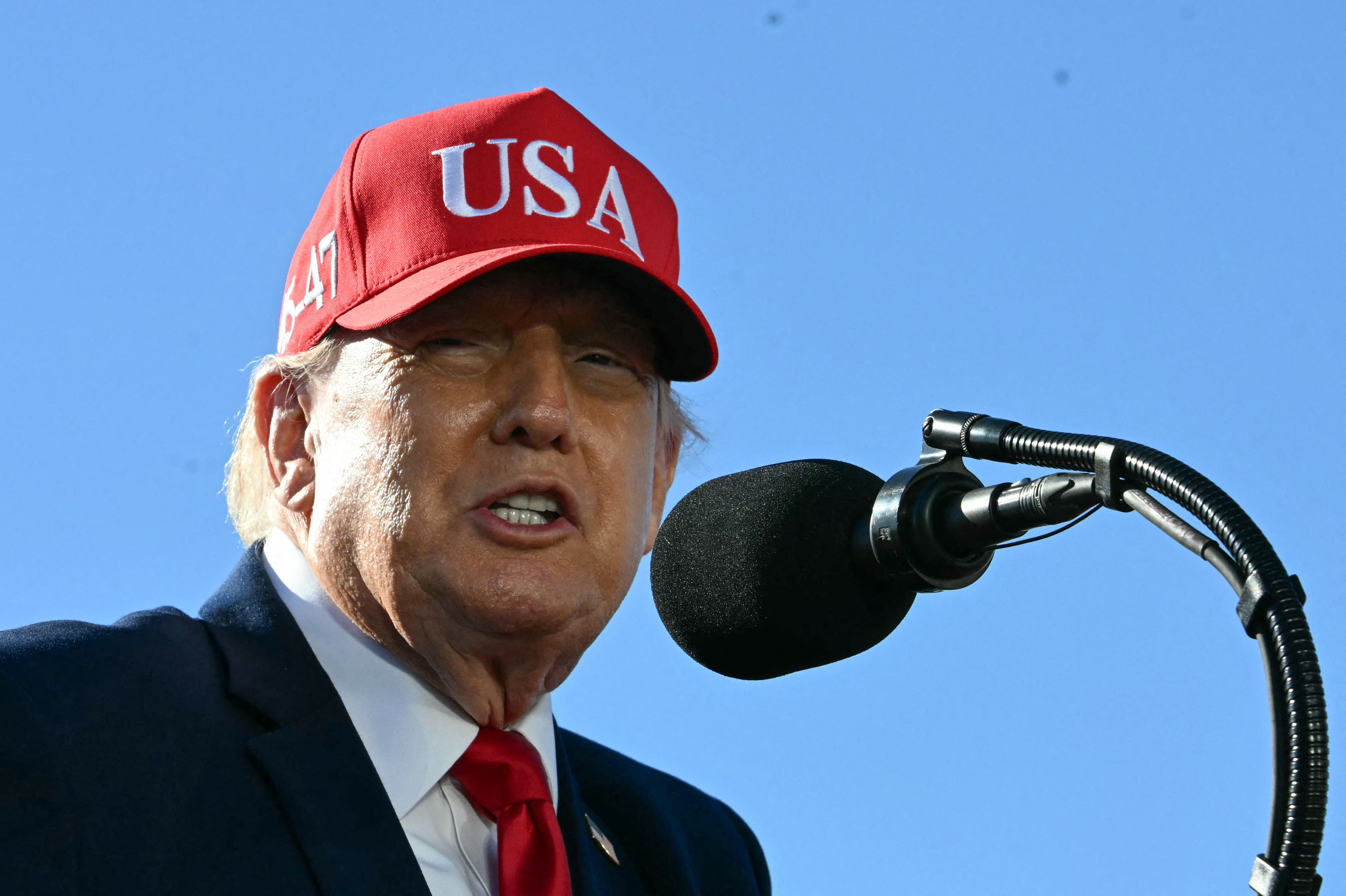 Mann mit roter "USA"-Kappe spricht ins Mikrofon vor blauem Himmel.