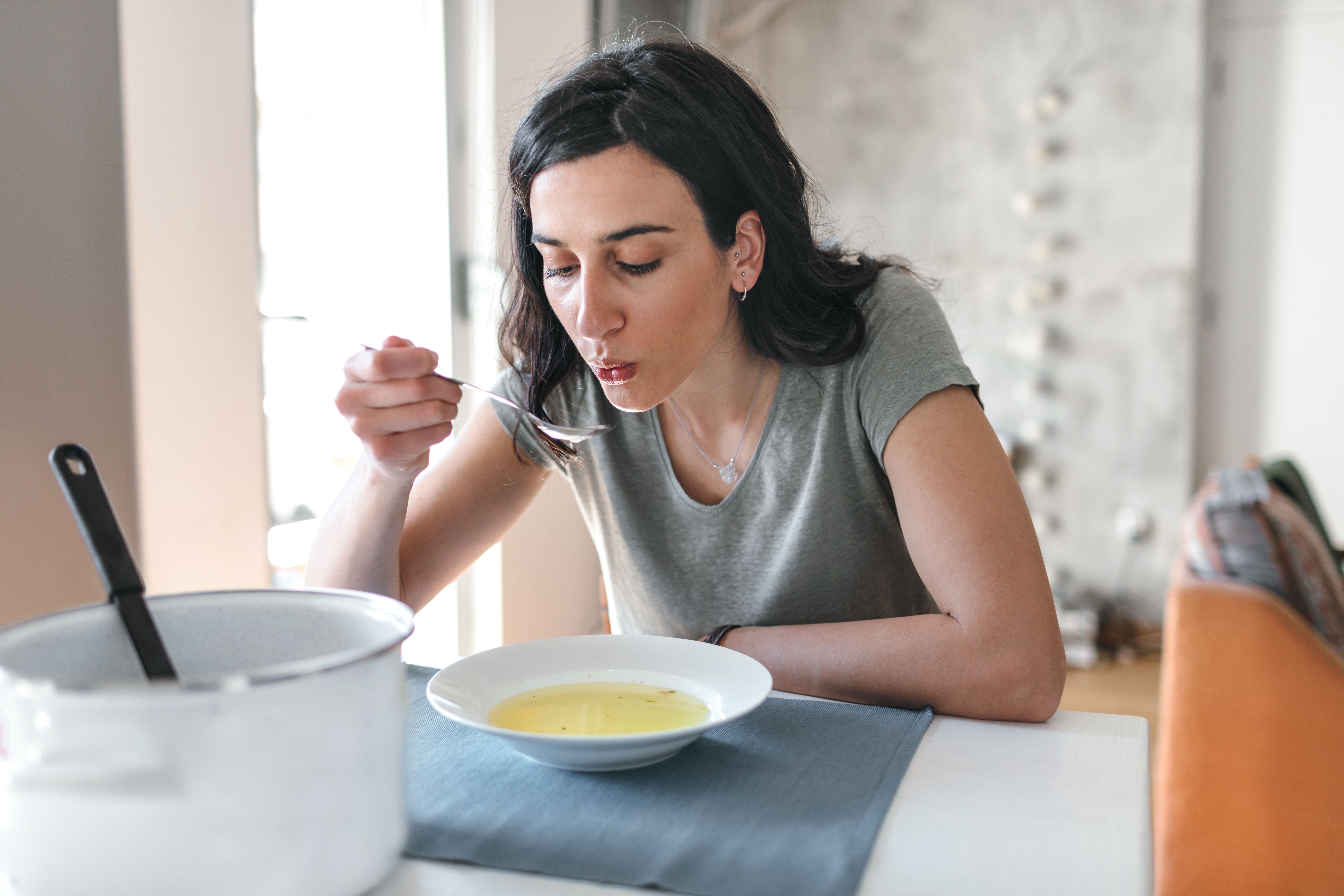 Junge Frau isst Suppe am Tisch, neben ihr steht ein Topf.