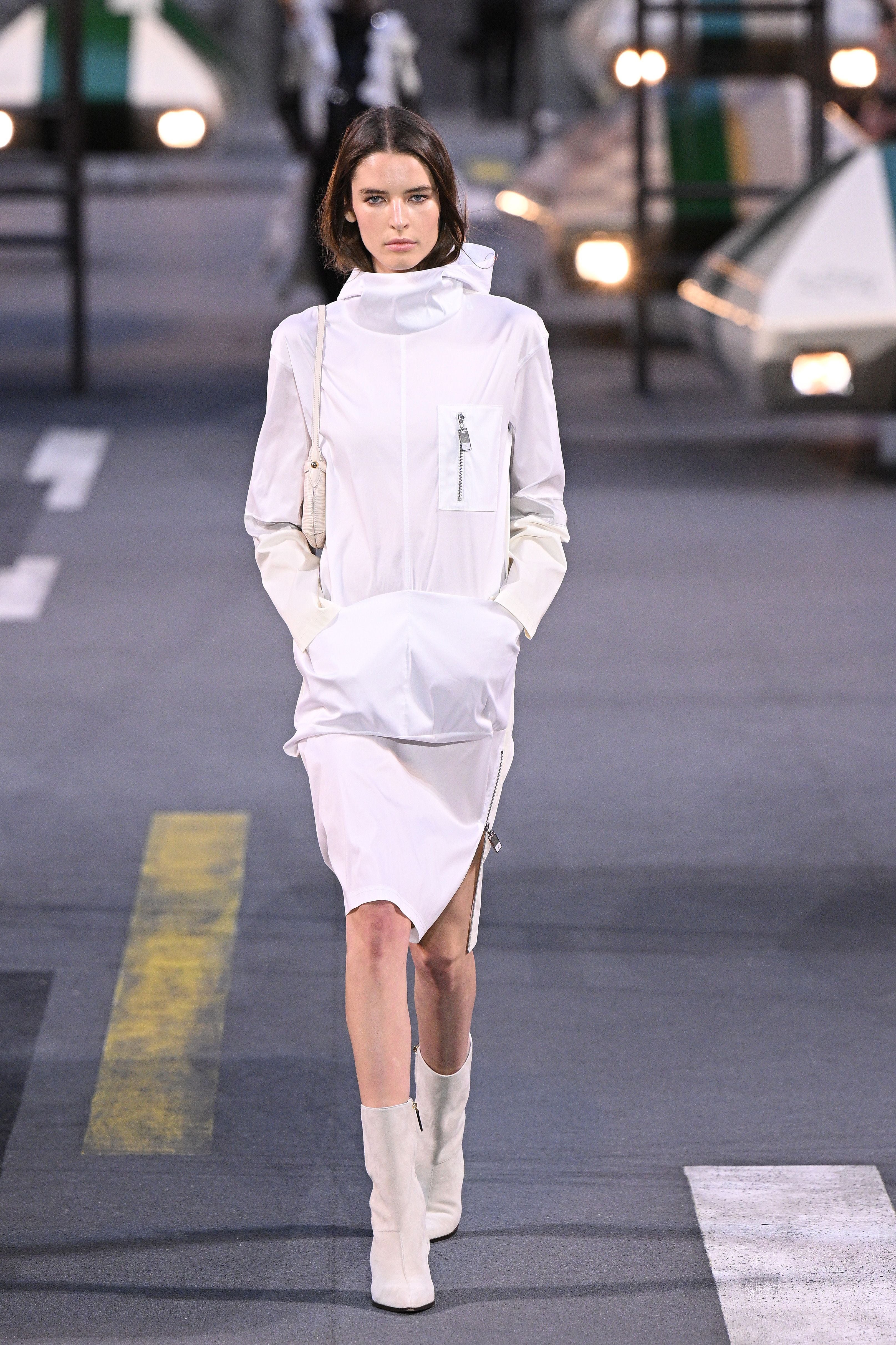 Model präsentiert weißes Kleid mit Stehkragen und Stiefeln auf dem Laufsteg bei einer Modenschau.