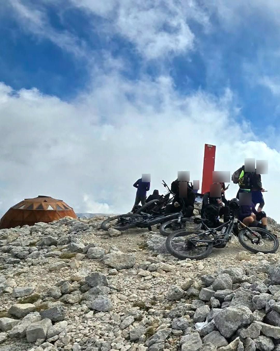 Gruppe mit Mountainbikes auf felsigem Gipfel bei einem roten Kreuz und einer Kuppelhütte in Italien.