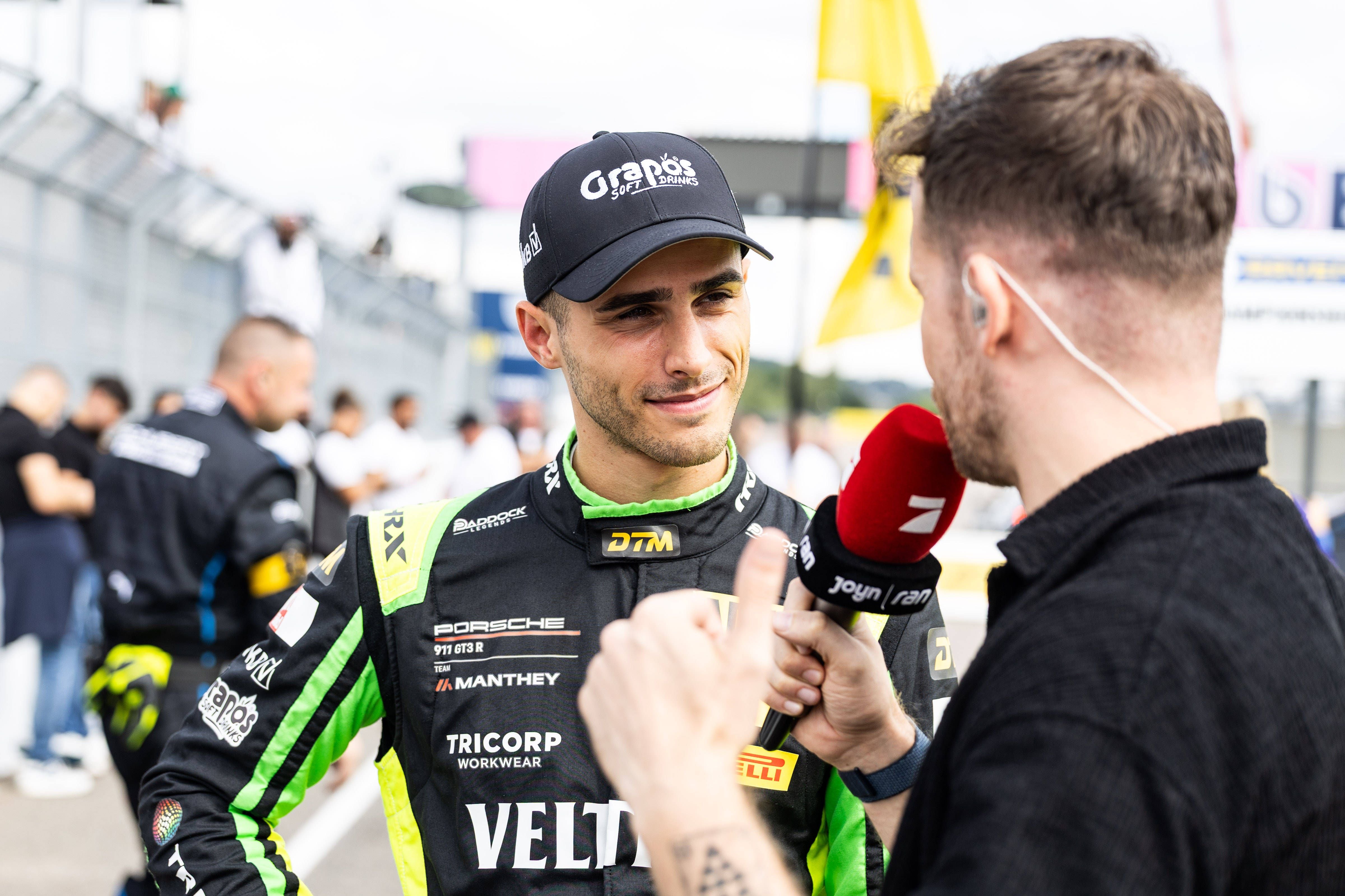 Thomas Preining lächelt im schwarzen Rennanzug mit Cap bei einem TV-Interview an der Rennstrecke.