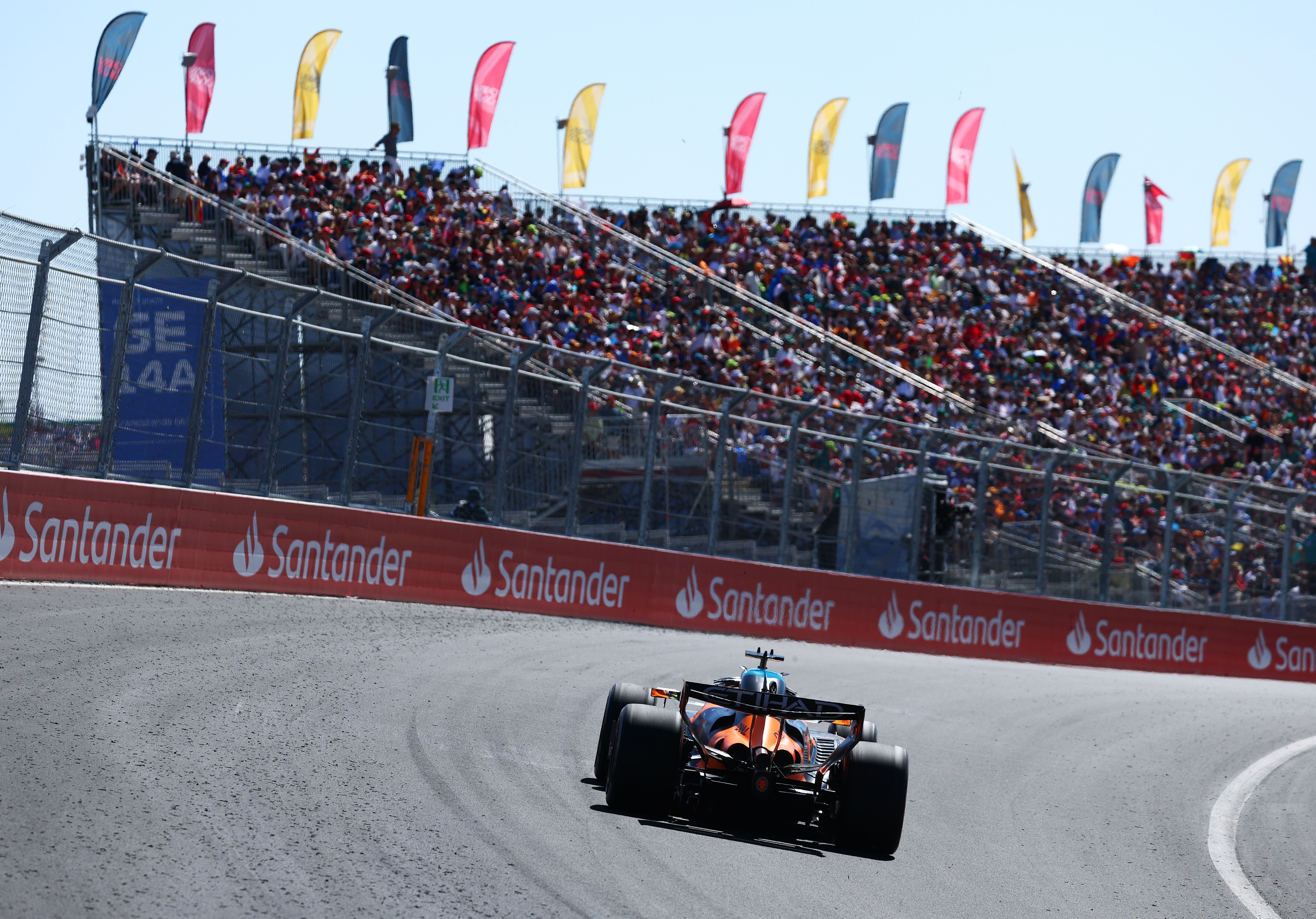 Ein orange-schwarzer McLaren-Rennwagen fährt bei Sonnenschein auf der Rennstrecke in eine Kurve vor gut gefüllten Zuschauerrängen.