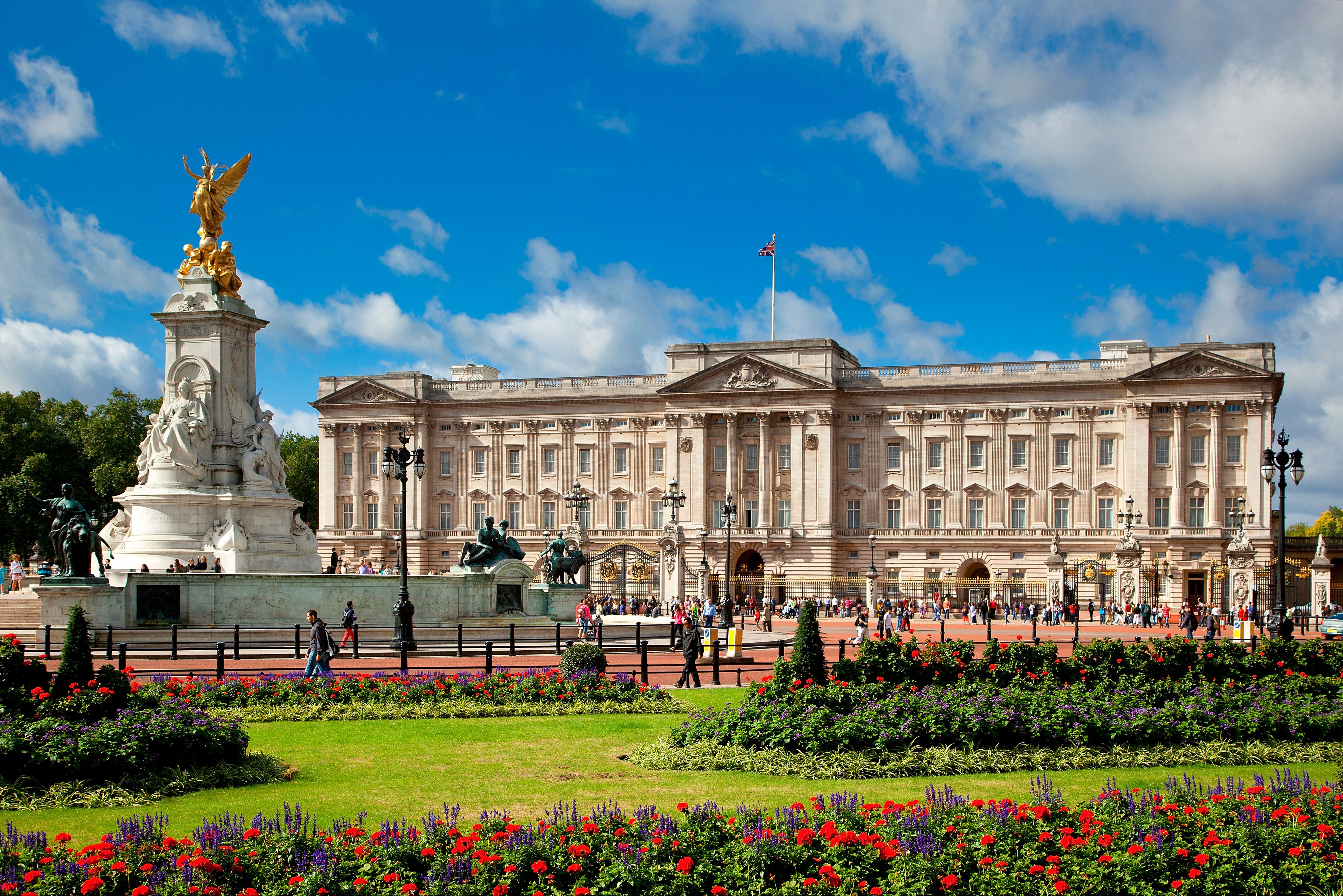 Buckingham Palace mit Blumenbeeten und Menschen vor dem Victoria Memorial an einem sonnigen Tag.
