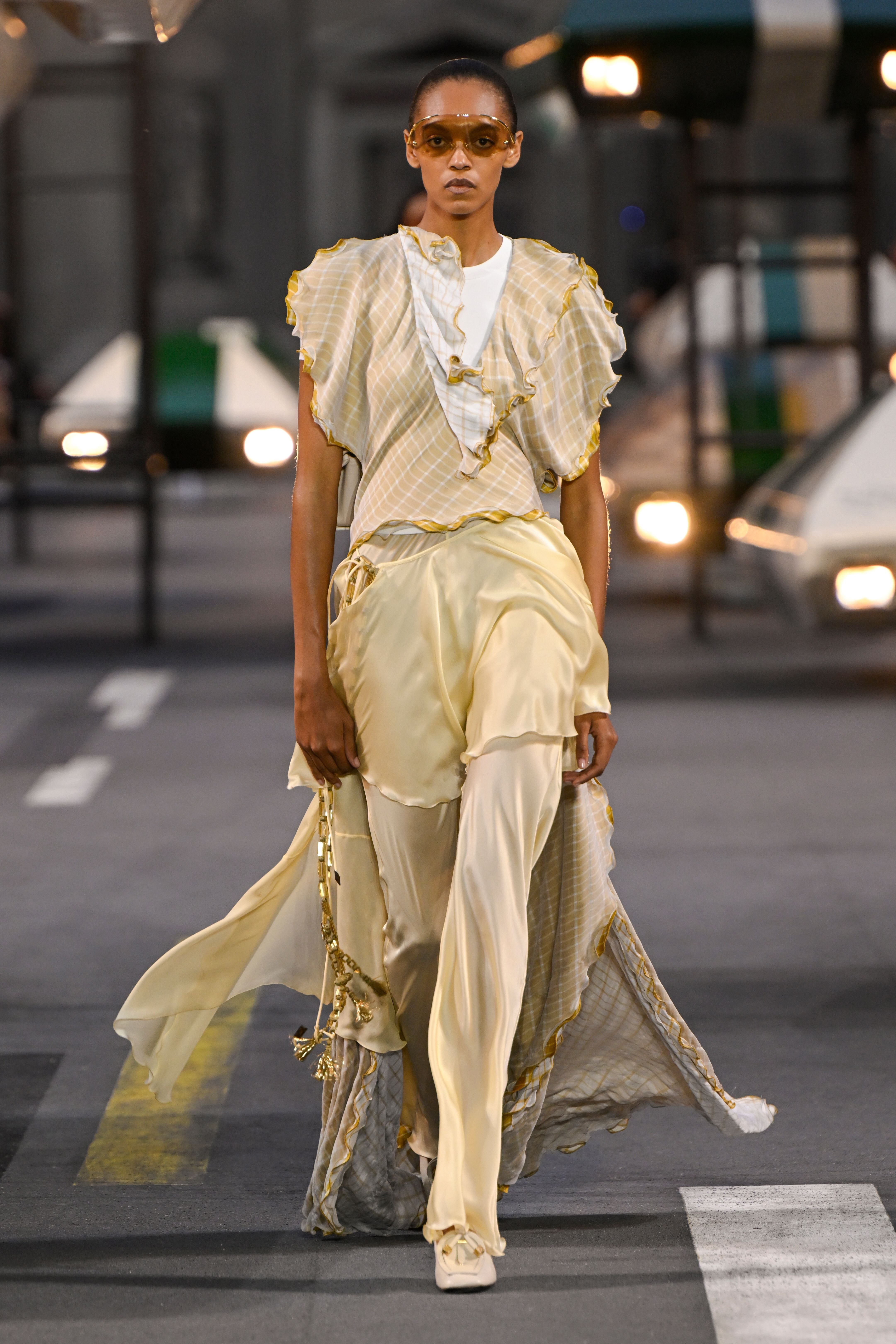 Model trägt fließendes, hellgelbes Outfit mit Rüschen und Sonnenbrille auf dem Laufsteg.