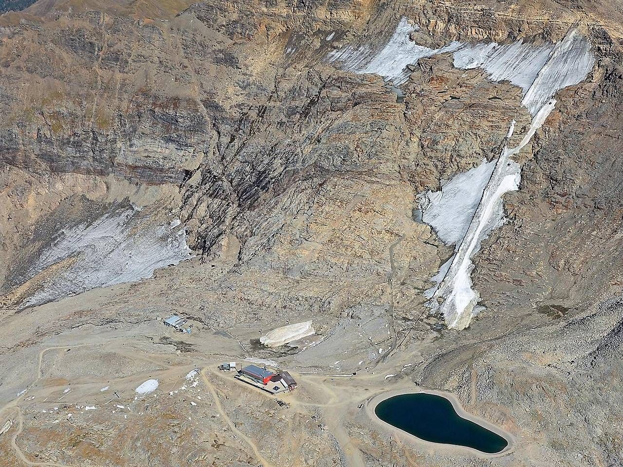 Berglandschaft mit Gletscherresten, Hütte und kleinem See, kaum Schnee sichtbar.