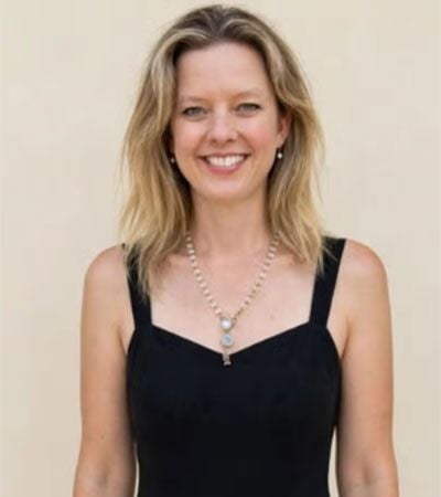 Frau mit blonden Haaren lächelt, trägt ein schwarzes Kleid und eine Perlenkette.