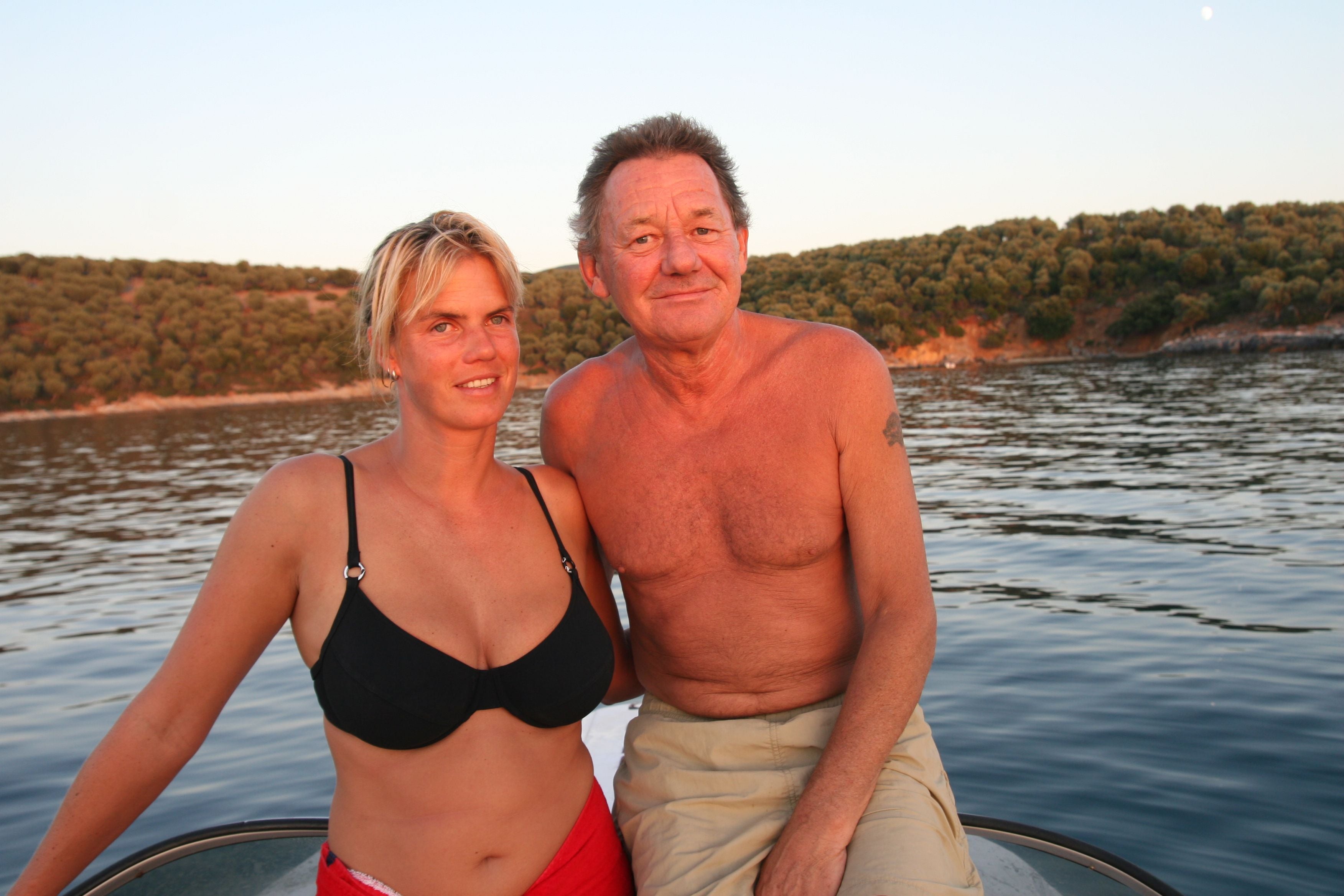 Ein Mann und eine Frau sitzen entspannt auf einem Boot vor bewaldeter Küste bei Sonnenuntergang.
