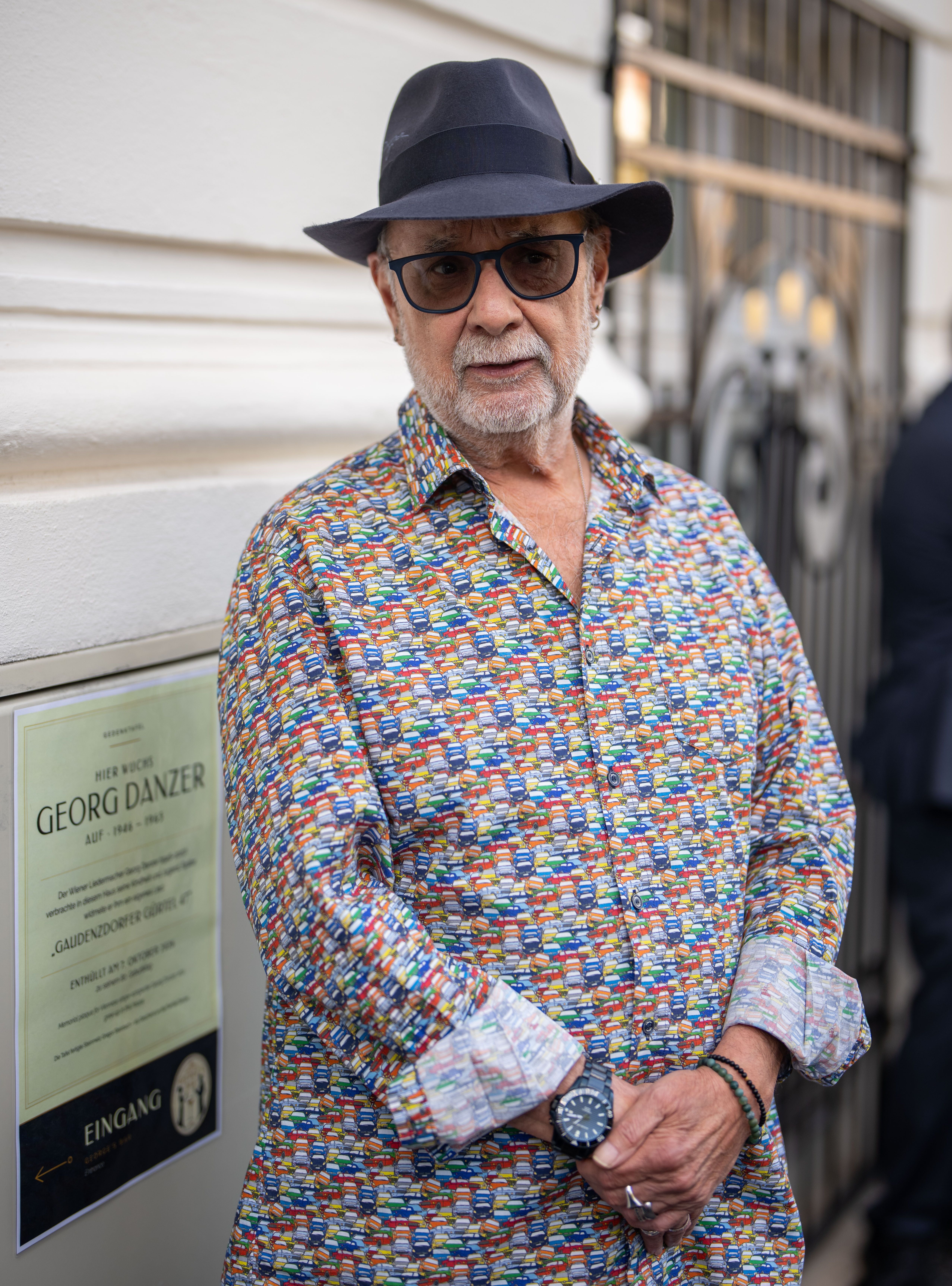 Älterer Mann mit Hut und bunter Brille steht vor einer Gedenktafel an einer Hauswand.