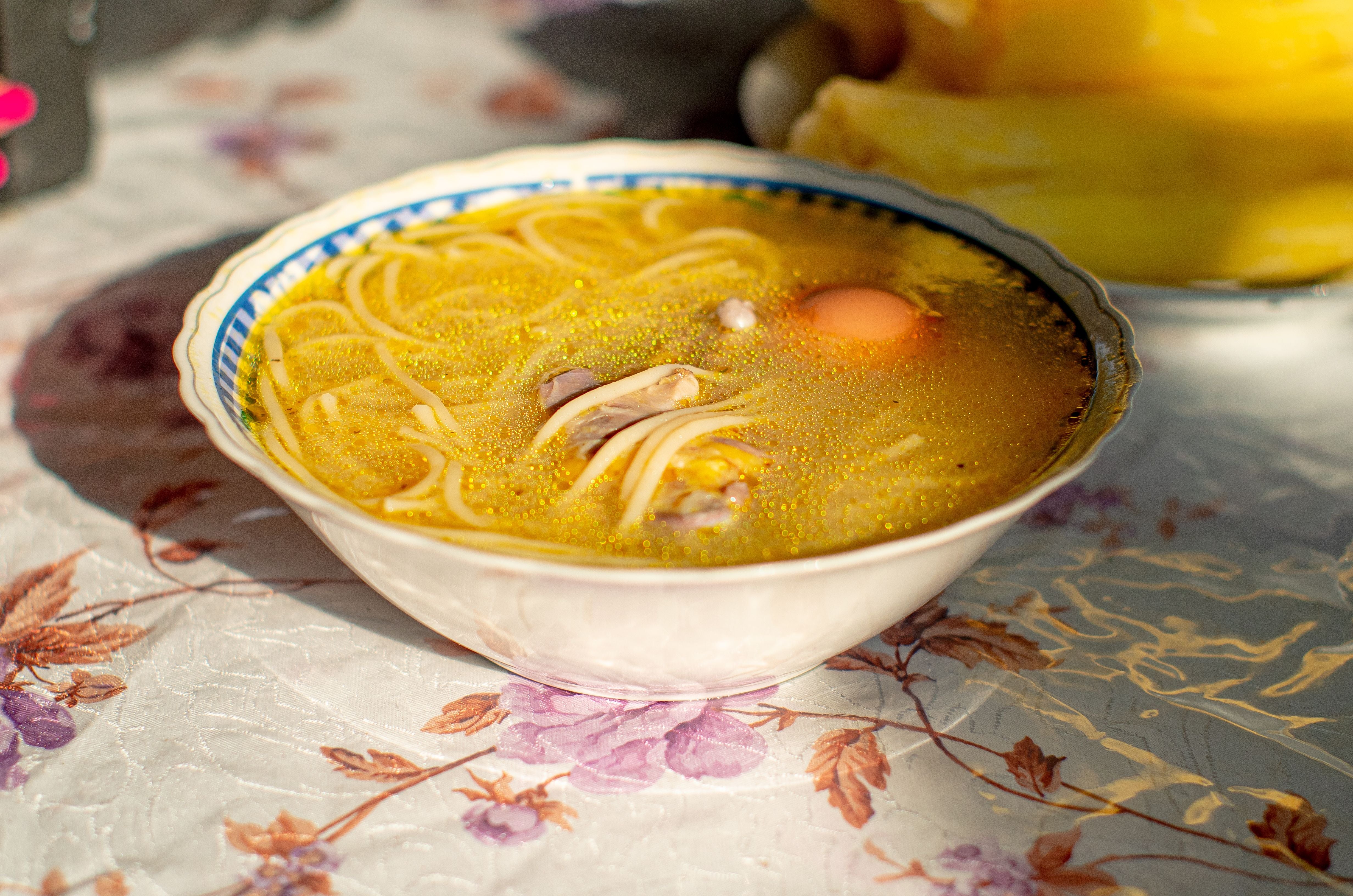 Schüssel mit Hühnersuppe, Nudeln und einem rohen Ei auf geblümter Tischdecke.