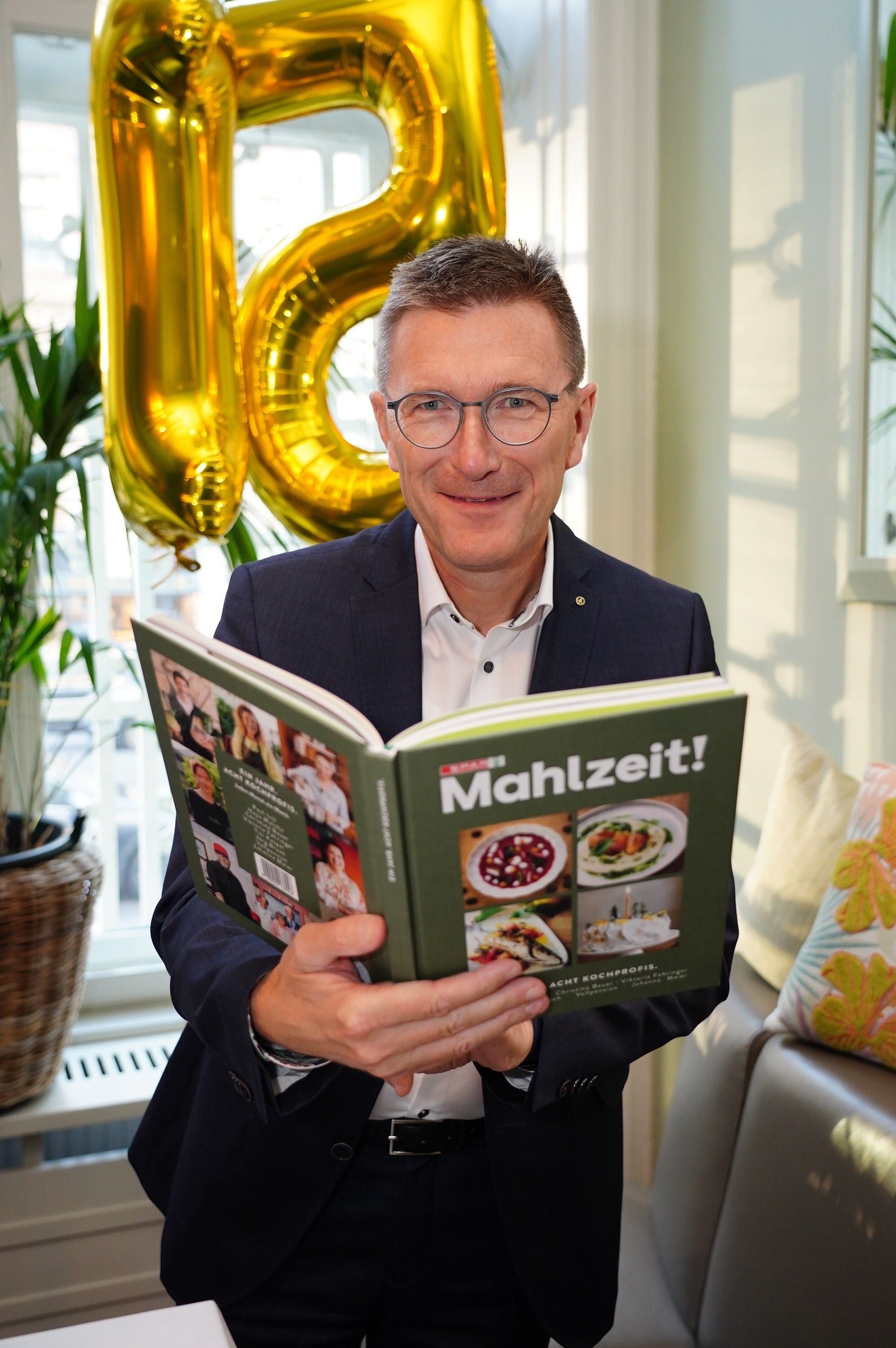 Mann mit Brille hält das Buch "Mahlzeit!" vor goldenen 15-Jahre-Ballon-Dekoration.