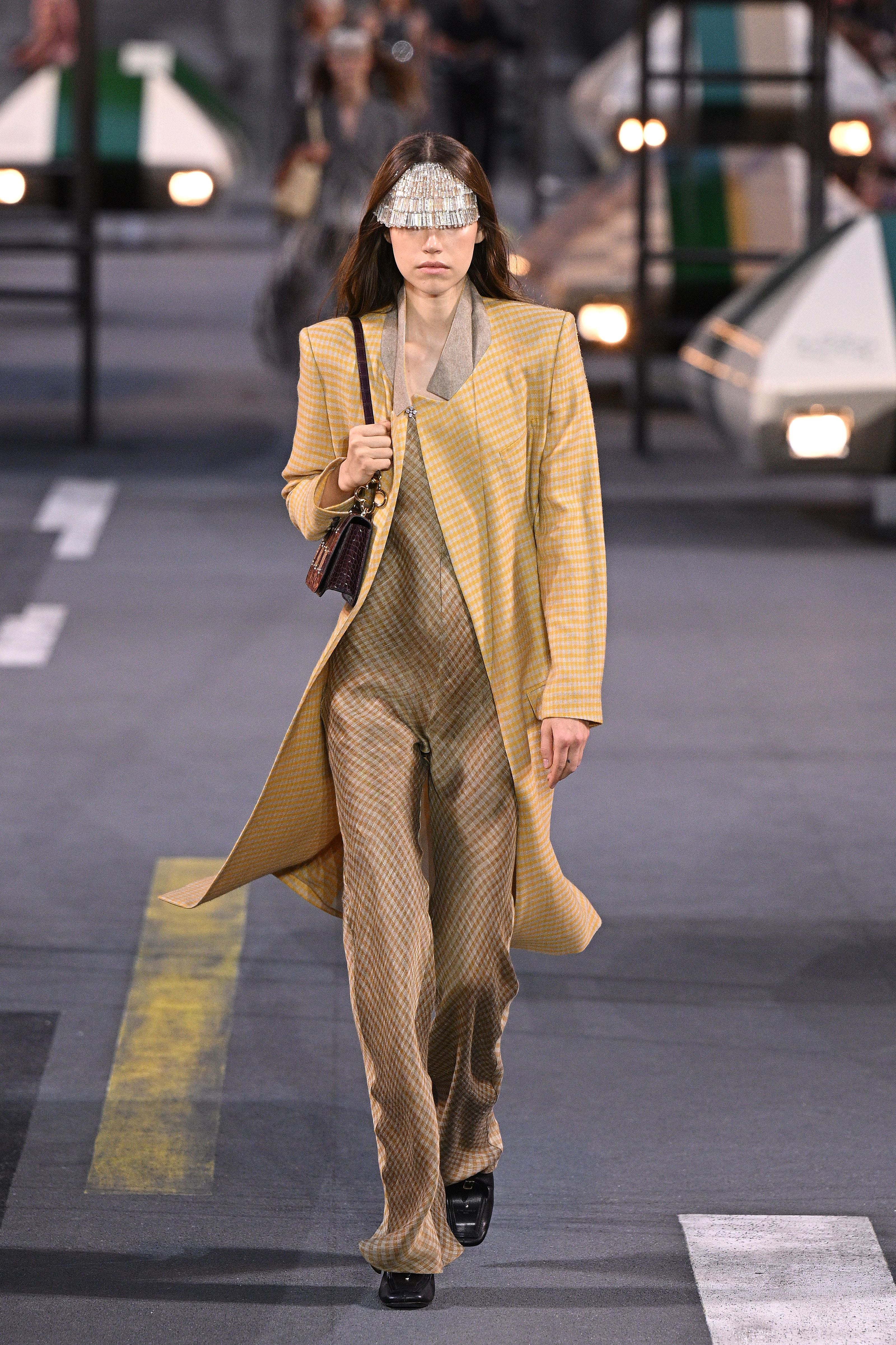 Model mit gelbem Karomantel, gemusterter Hose und silberner Augenmaske auf dem Laufsteg.