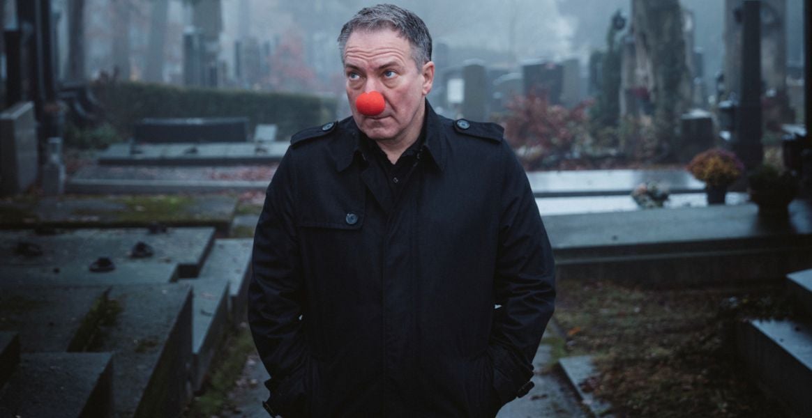 Ein Mann mit roter Clownnase steht in dunkler Kleidung auf einem nebligen Friedhof.