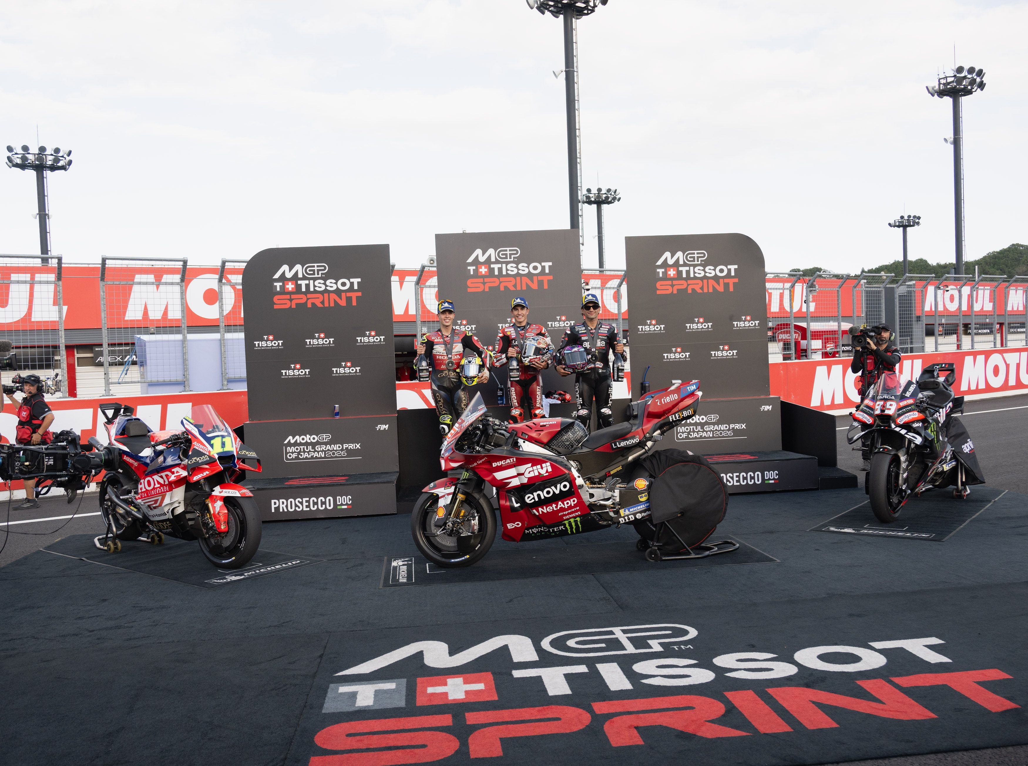 Drei MotoGP-Fahrer stehen auf den Podestplätzen vor Werbewänden der Tissot-Sprint-Serie, davor parken drei Rennmotorräder.