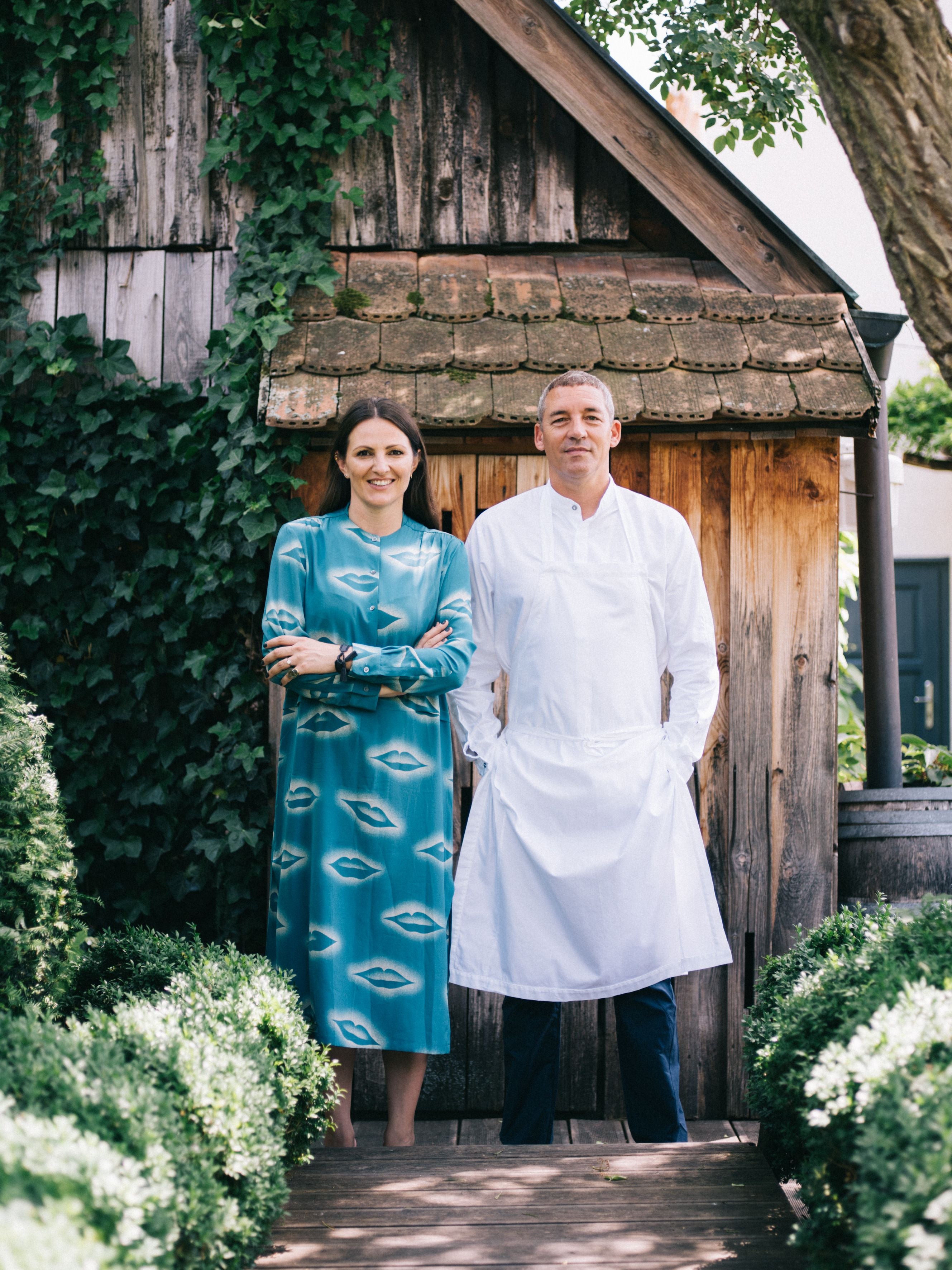 Eine Frau im blauen Kleid und ein Mann in Kochkleidung stehen vor einem Holzhaus im Grünen.