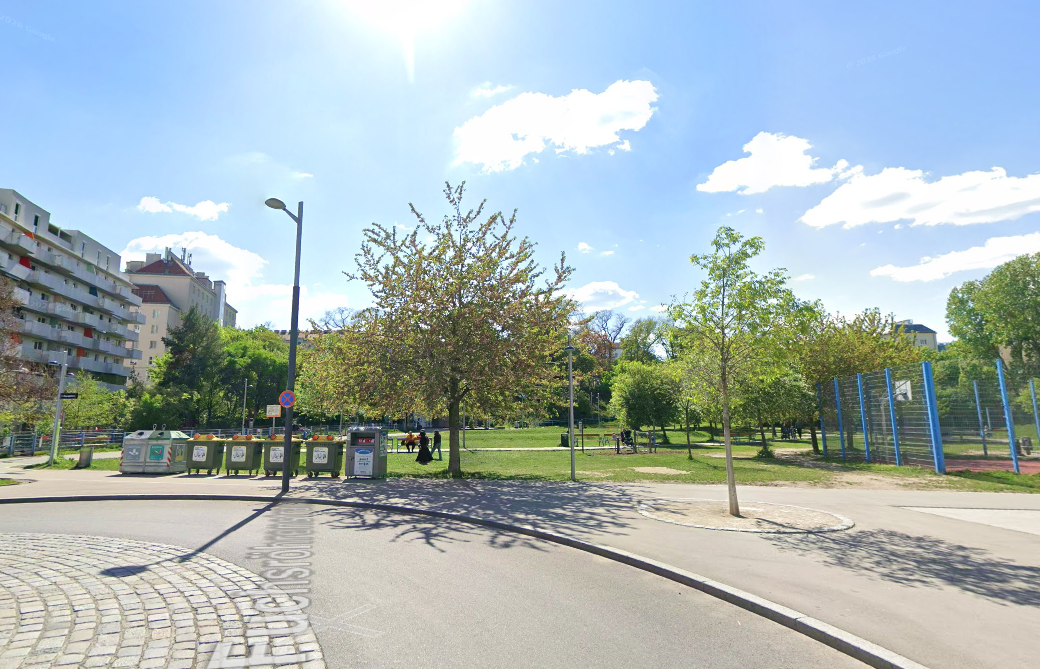 Sonniger Park mit Bäumen, Gehwegen, Sitzbänken und daneben einem Spiel- oder Sportplatz.