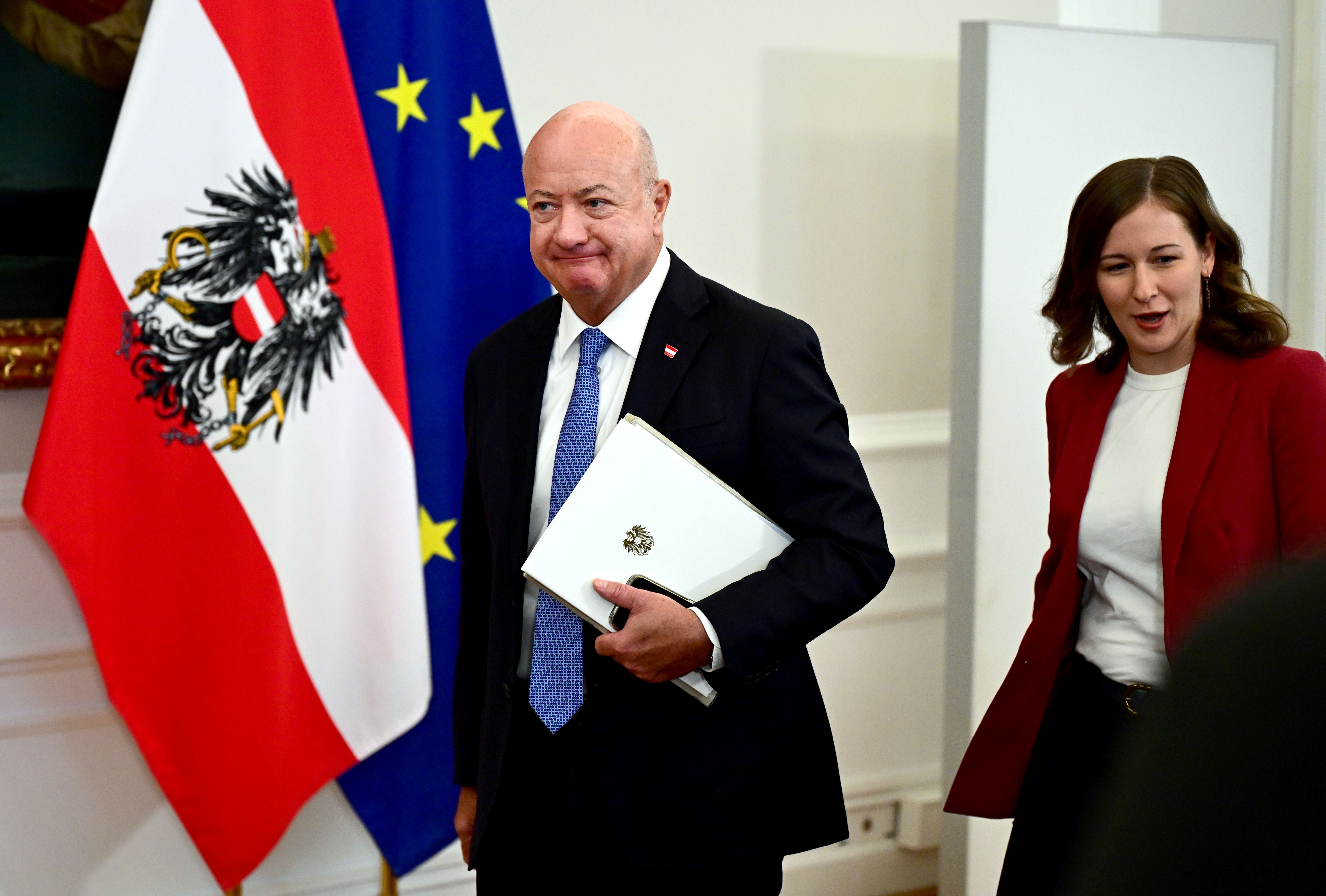 Mann im Anzug und Frau im Blazer gehen vor österreichischer und EU-Flagge in einem Raum.