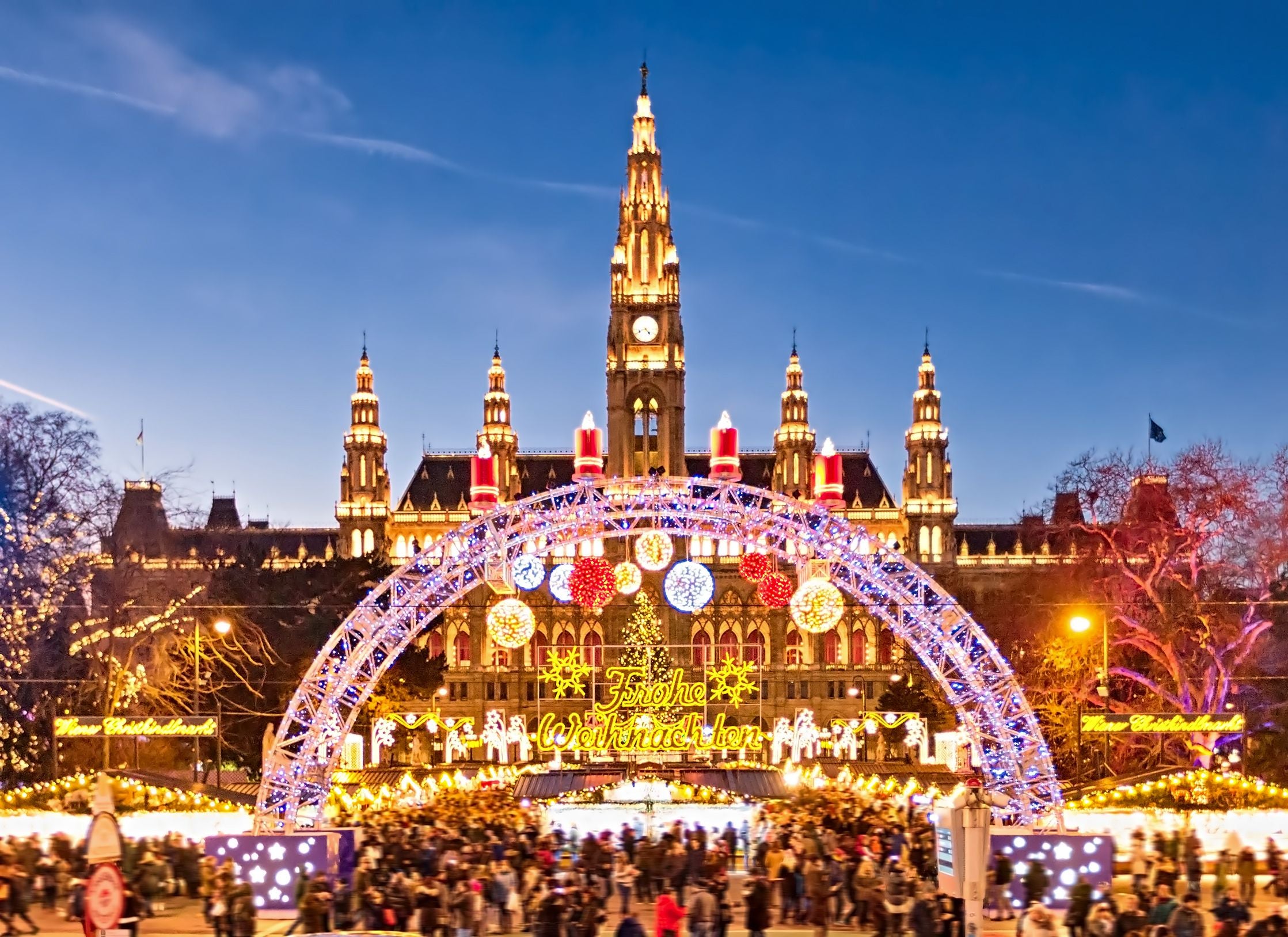 Blick auf den beleuchteten Wiener Weihnachtsmarkt am Rathausplatz mit „Frohe Weihnachten“-Schriftzug.